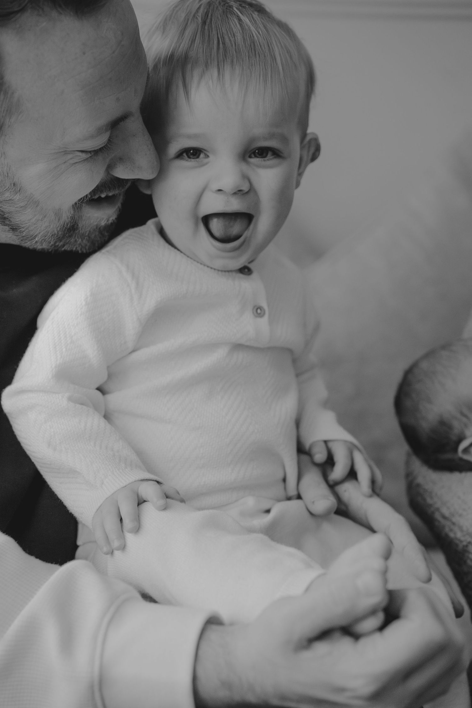 Black and white photo of a laughing baby in white onesie being held by a smiling father.