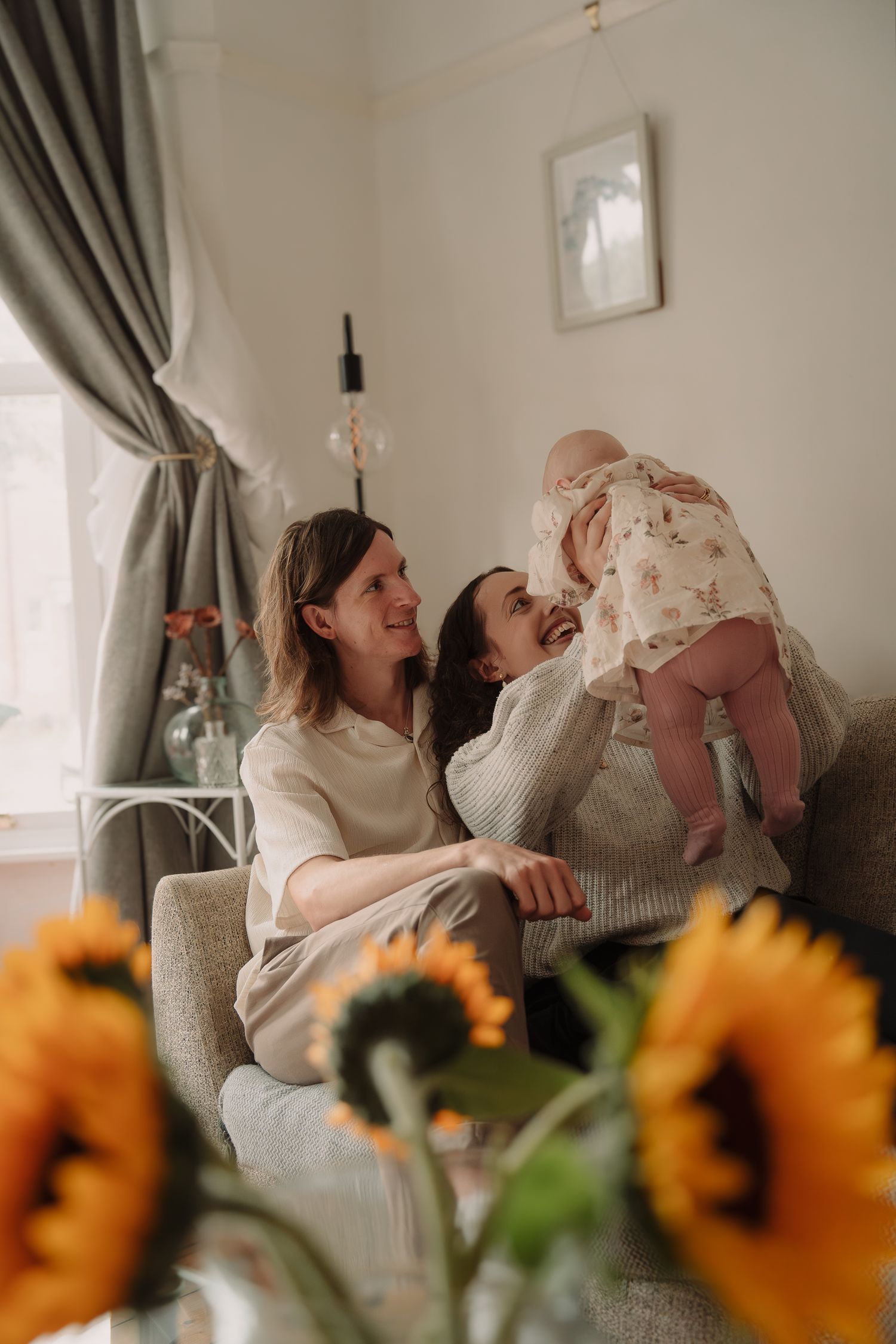 Joyful moment in living room with sunflowers in foreground while family shares laughter on cozy gray armchair.