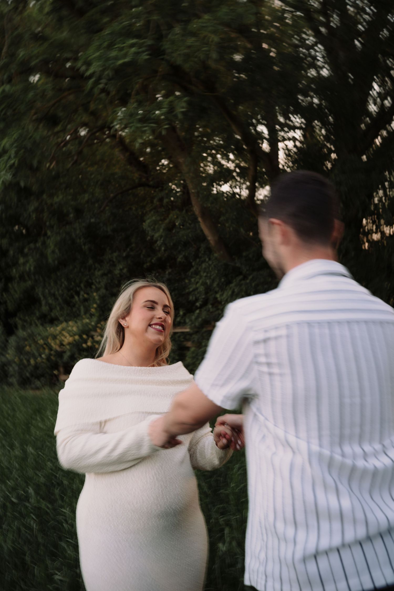Couple dancing outdoors in garden, woman in white dress smiling joyfully at man in striped shirt.