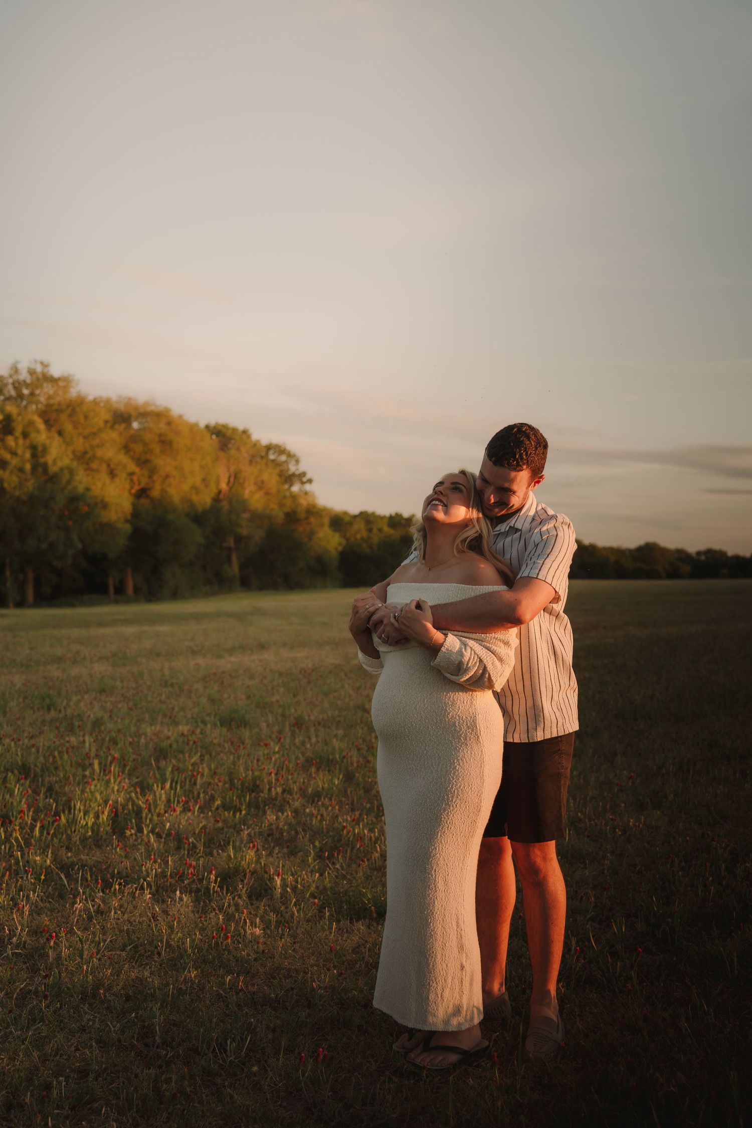 Couple embracing in golden hour sunlight in a green field, pregnant woman in white dress held by man in striped shirt.