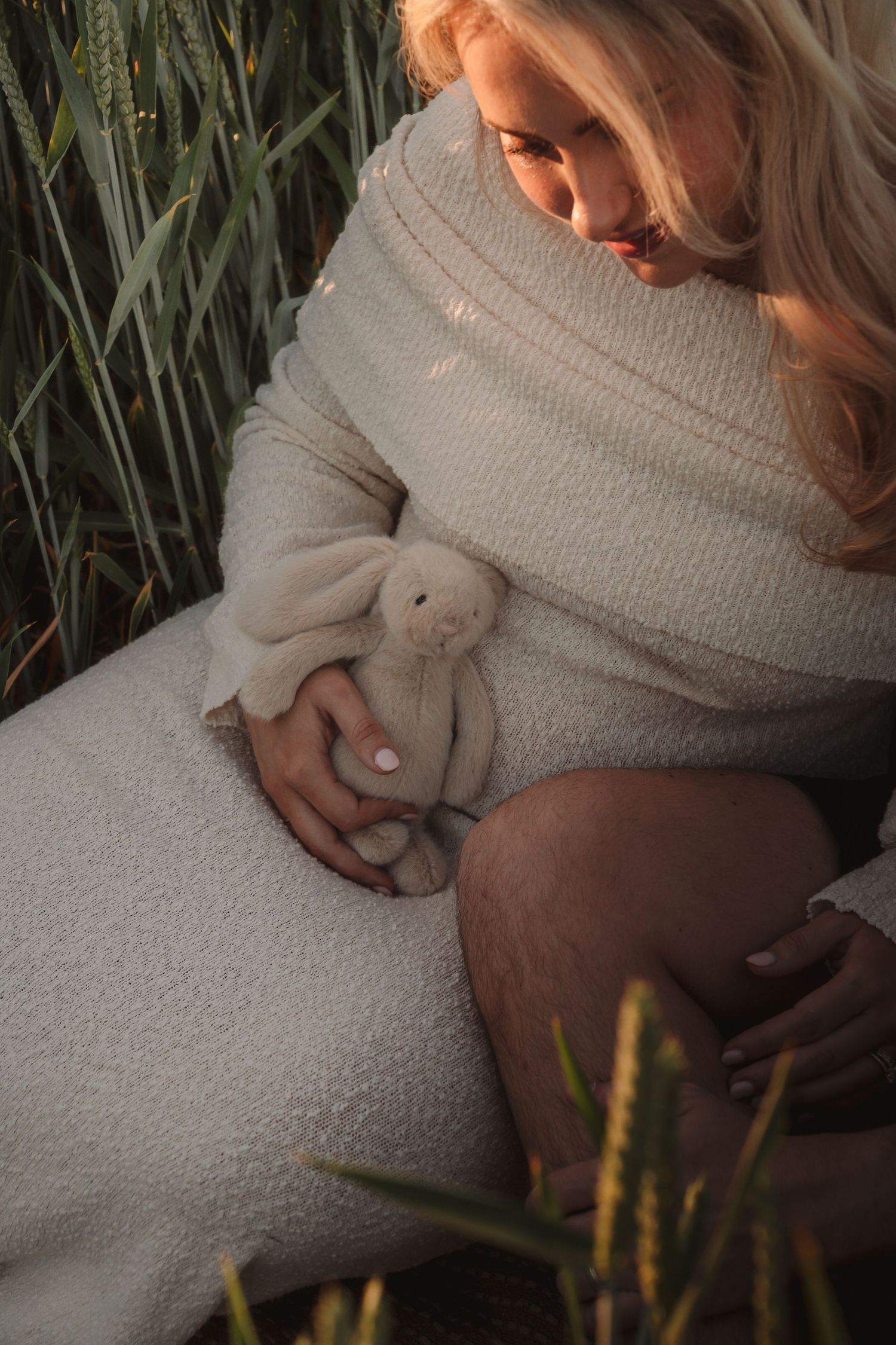 Pregnant woman in white knit dress holding a stuffed bunny toy while sitting among tall green wheat stalks at sunset.
