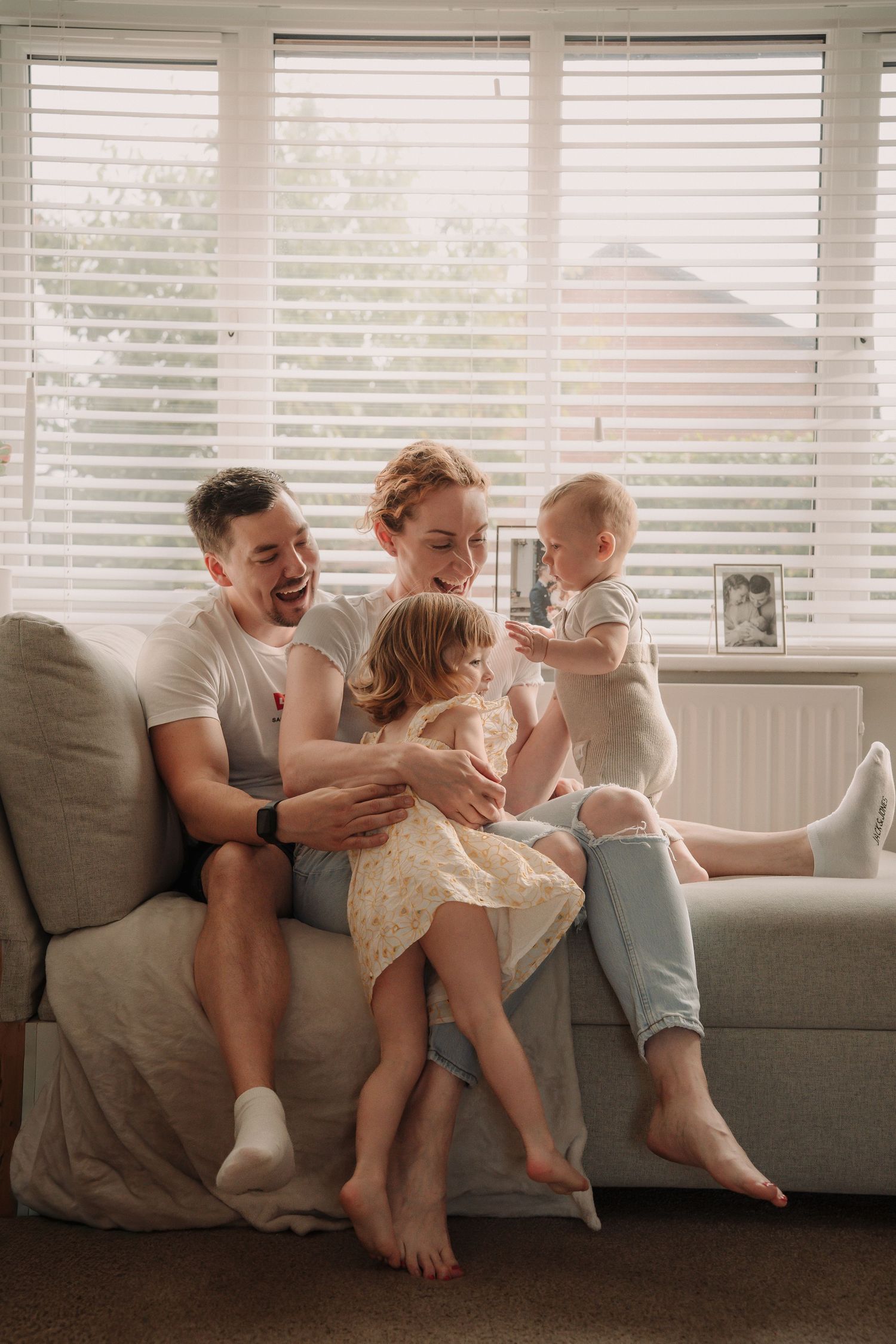 Family shares joyful moments together on a gray couch by a window with white blinds.