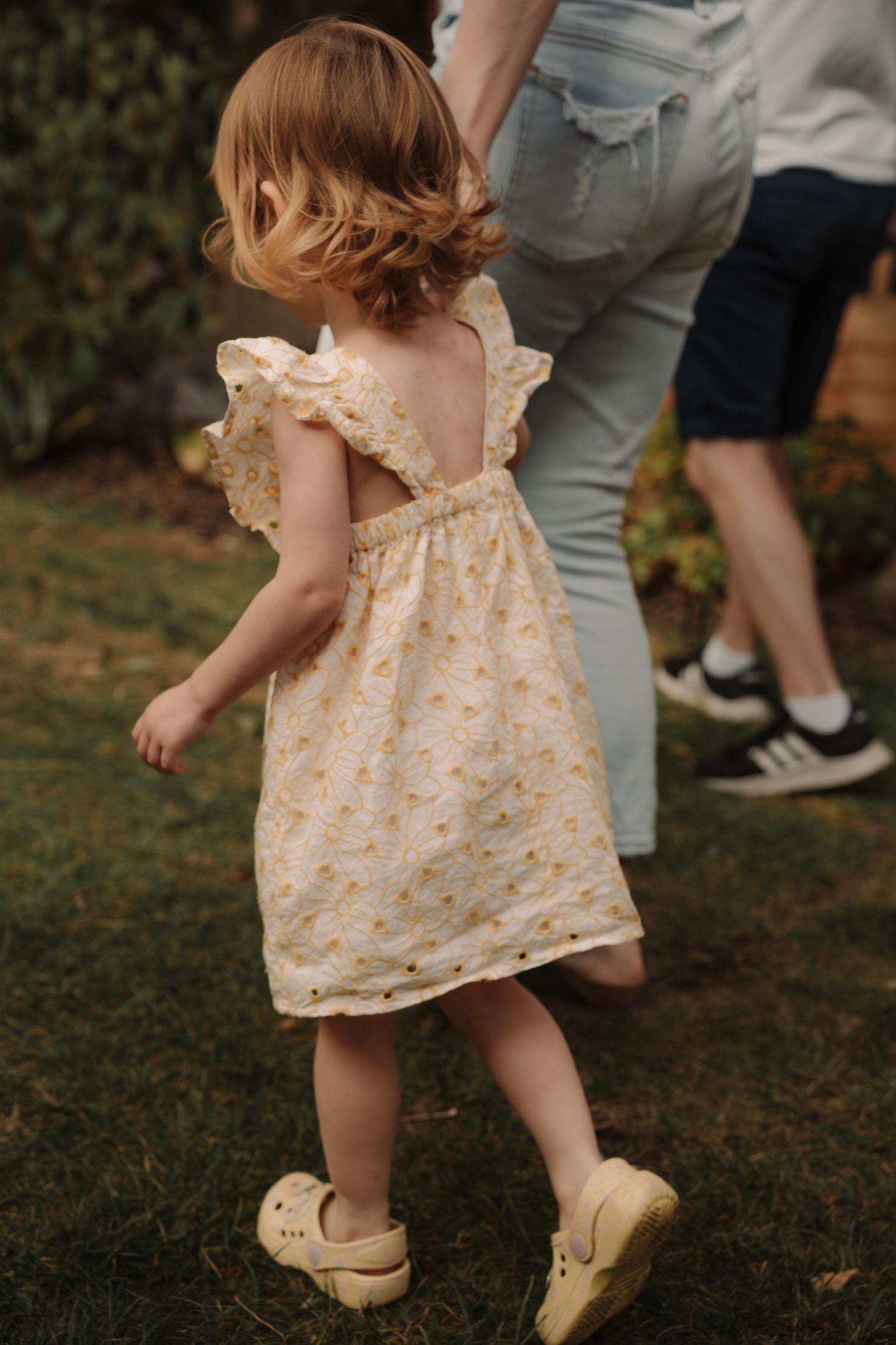 A small child in a floral dress and beige shoes walks on grass in a series of progressive steps.