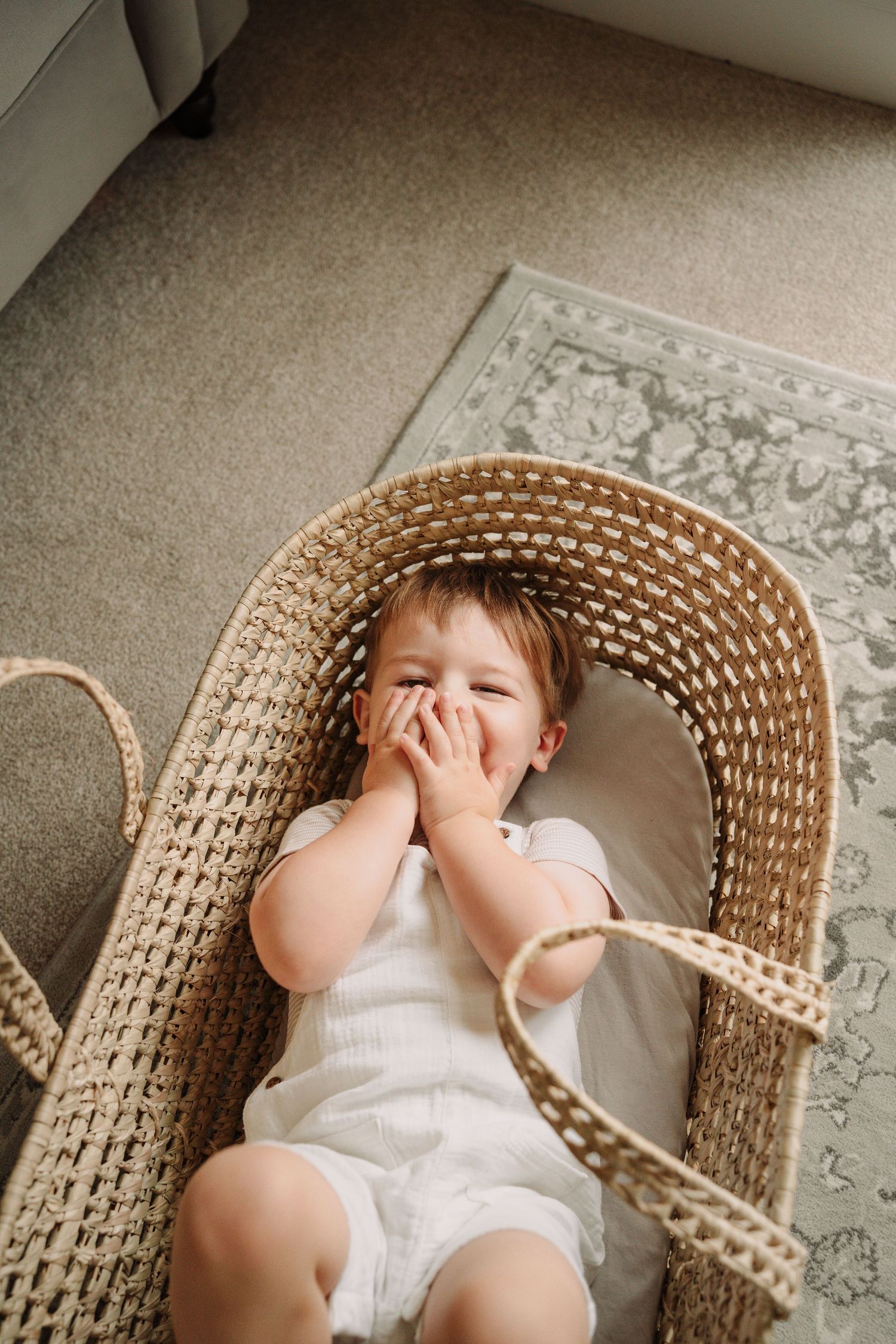 Child playing and laughing in wicker chair on patterned rug.