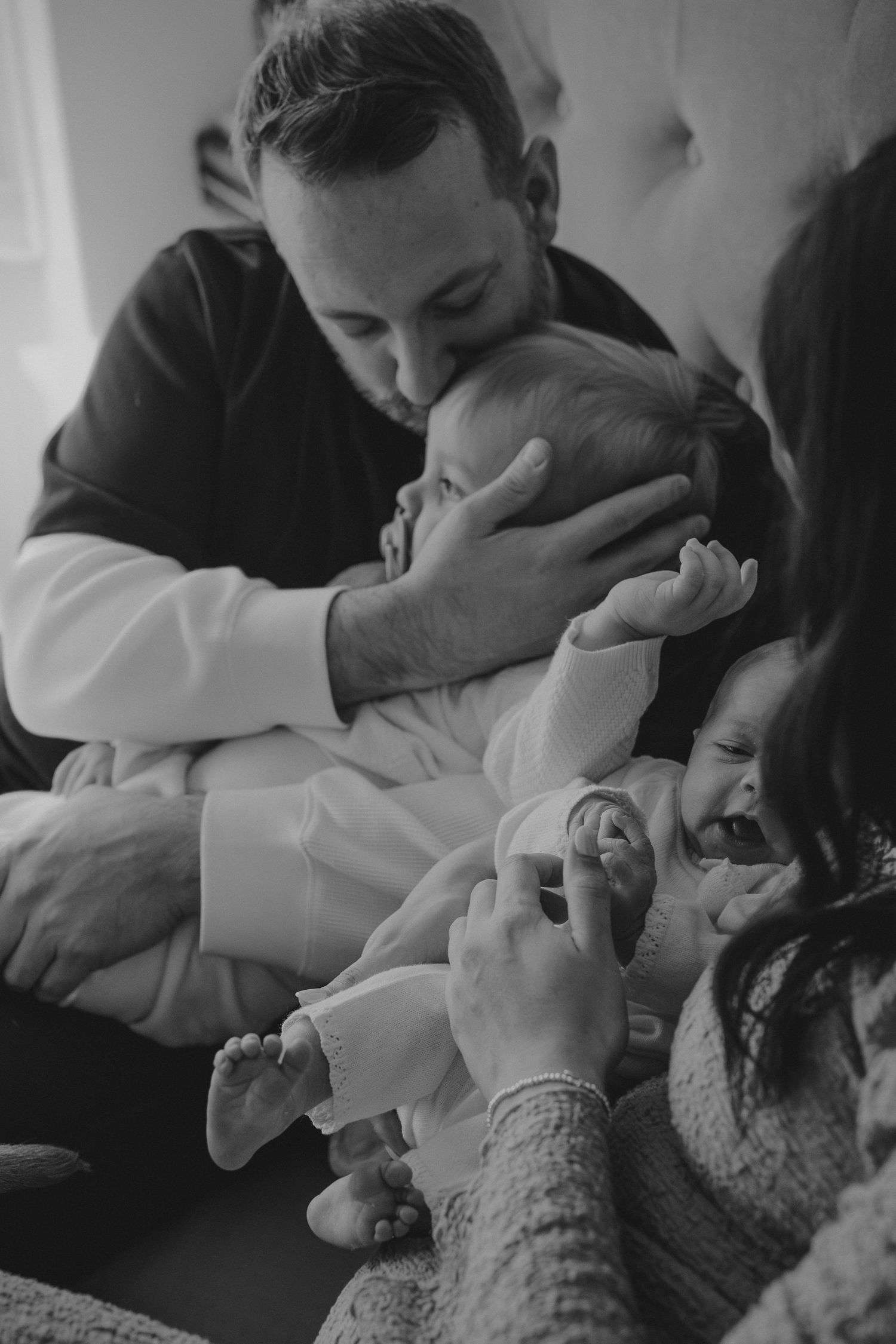 Black and white photo of father kissing newborn baby held with toddler sibling.