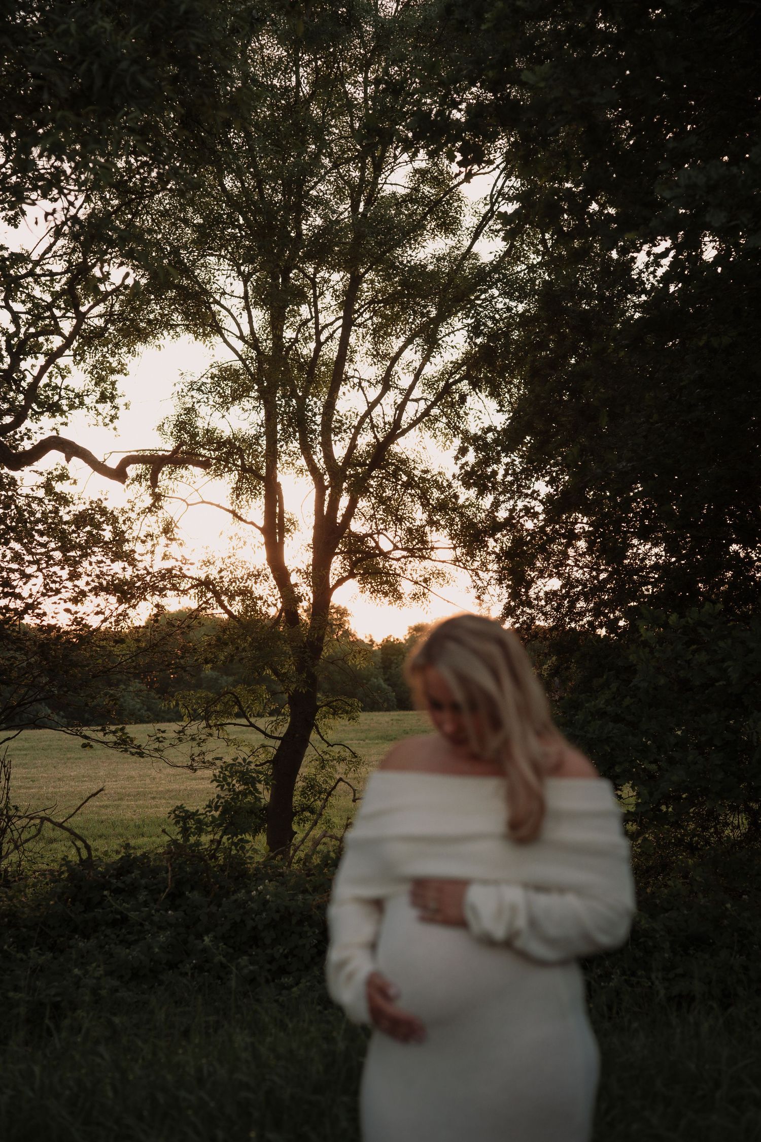 Pregnant woman in white off-shoulder dress cradling her bump at golden hour, surrounded by lush trees in a serene outdoor setting.