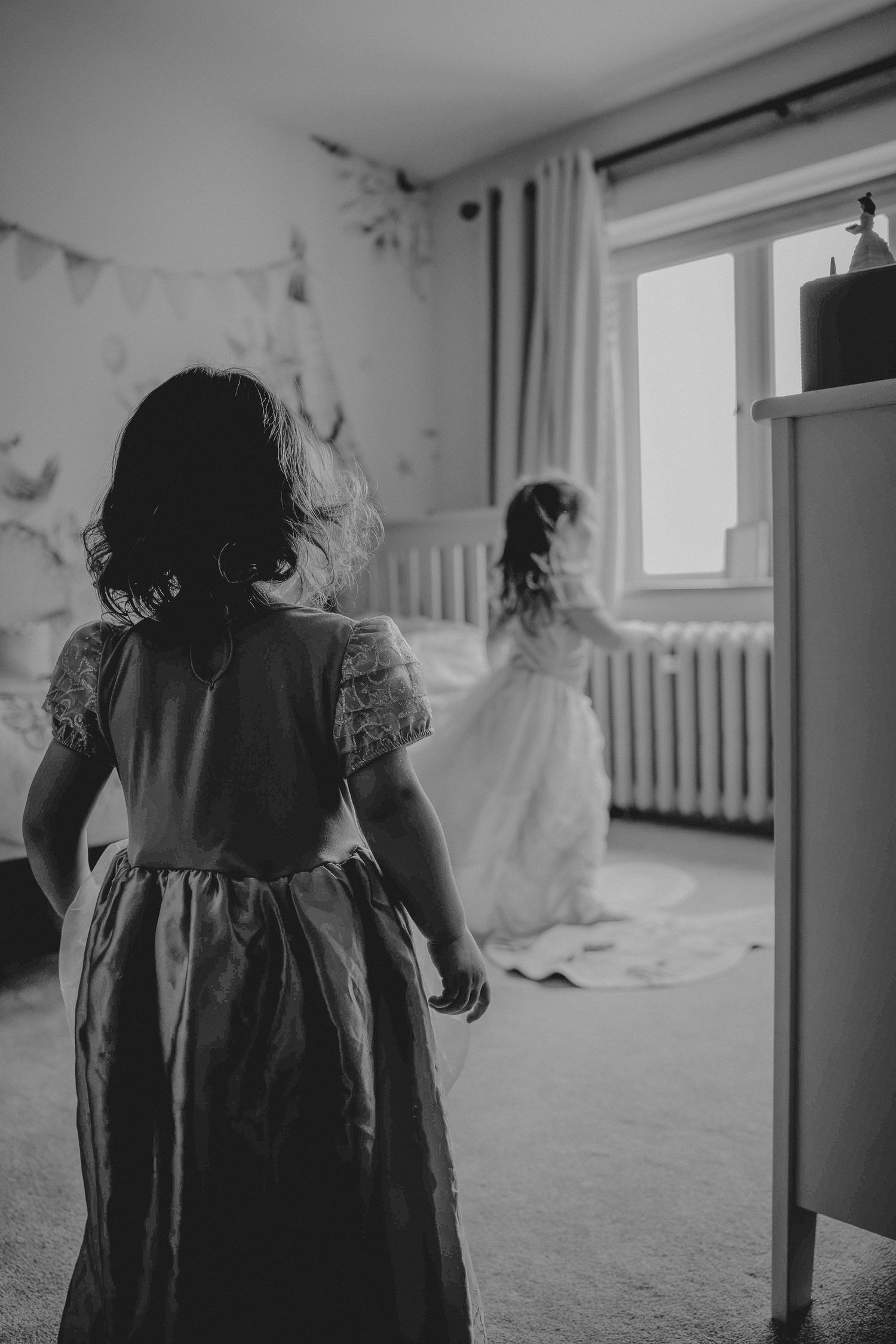 Black and white photo of two young children in dresses playing in a sunlit room with white curtains and a window.