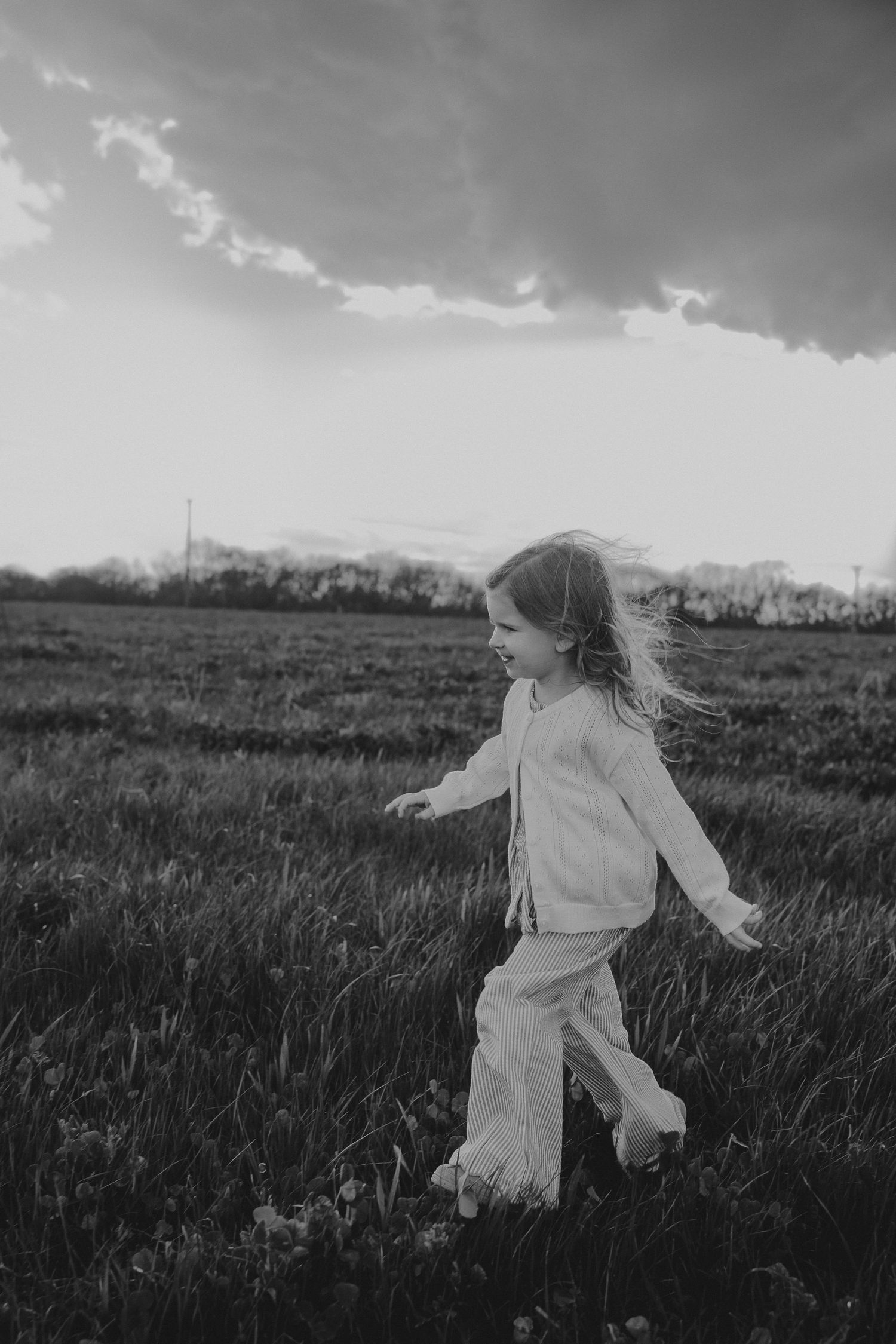 Black and white photo of a young girl in white outfit running through a grassy field under dramatic clouds.
