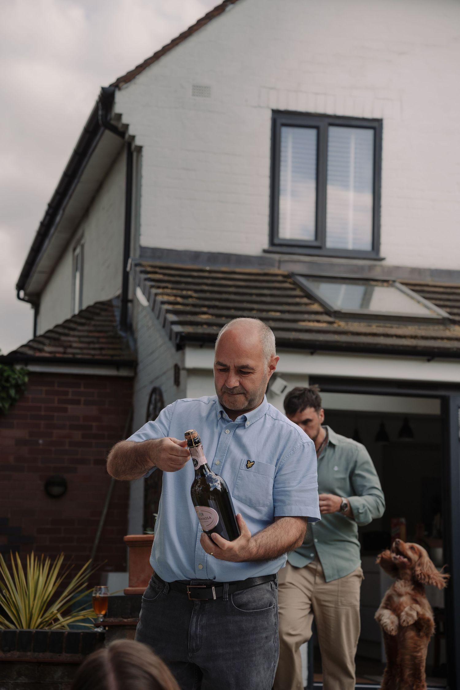 A bald man opens a champagne bottle outdoors near a house, with a man and a dog visible in the background.