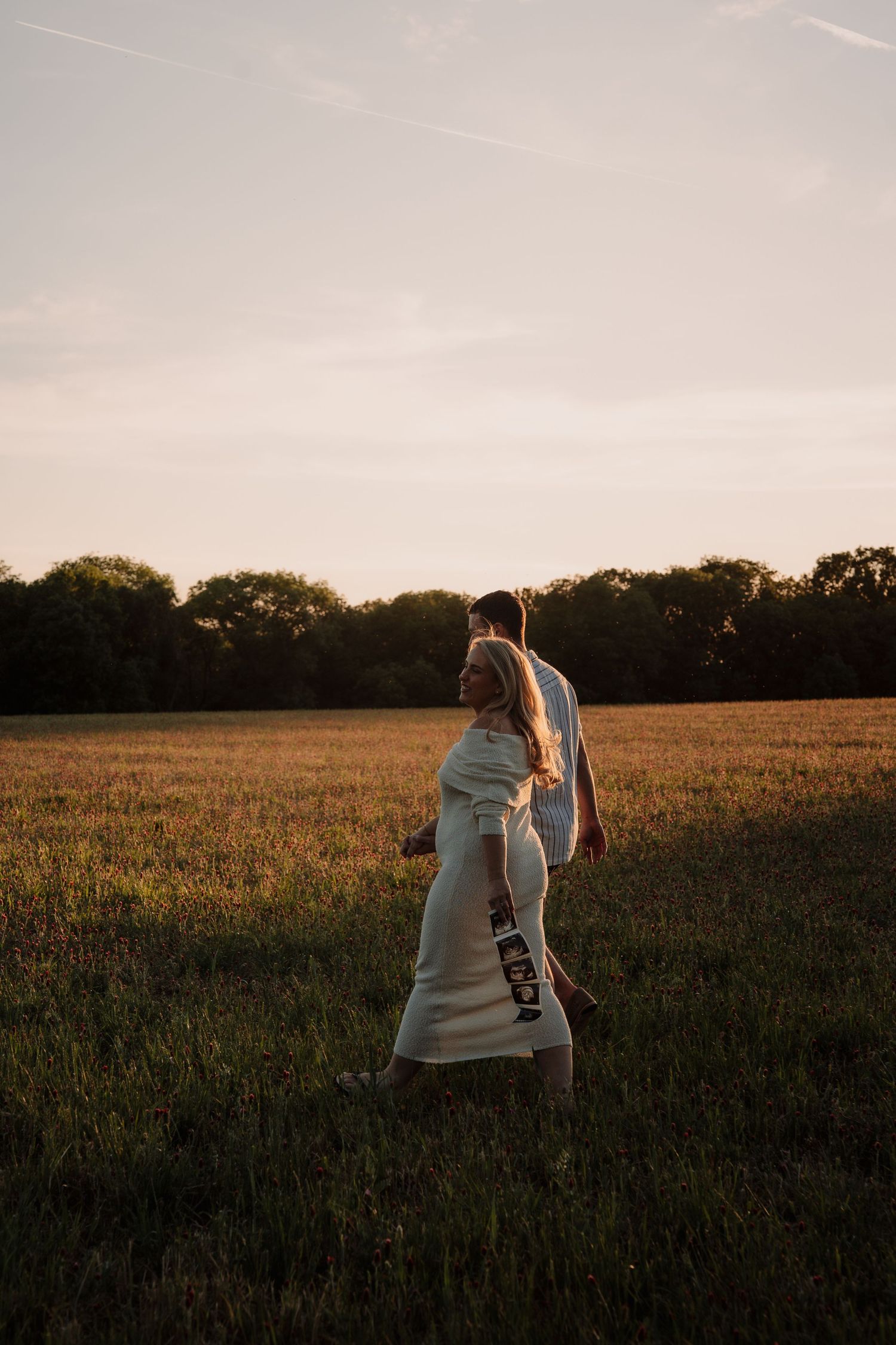 Couple walking in golden sunset field, woman in white dress with flowing veil, romantic outdoor maternity photo.