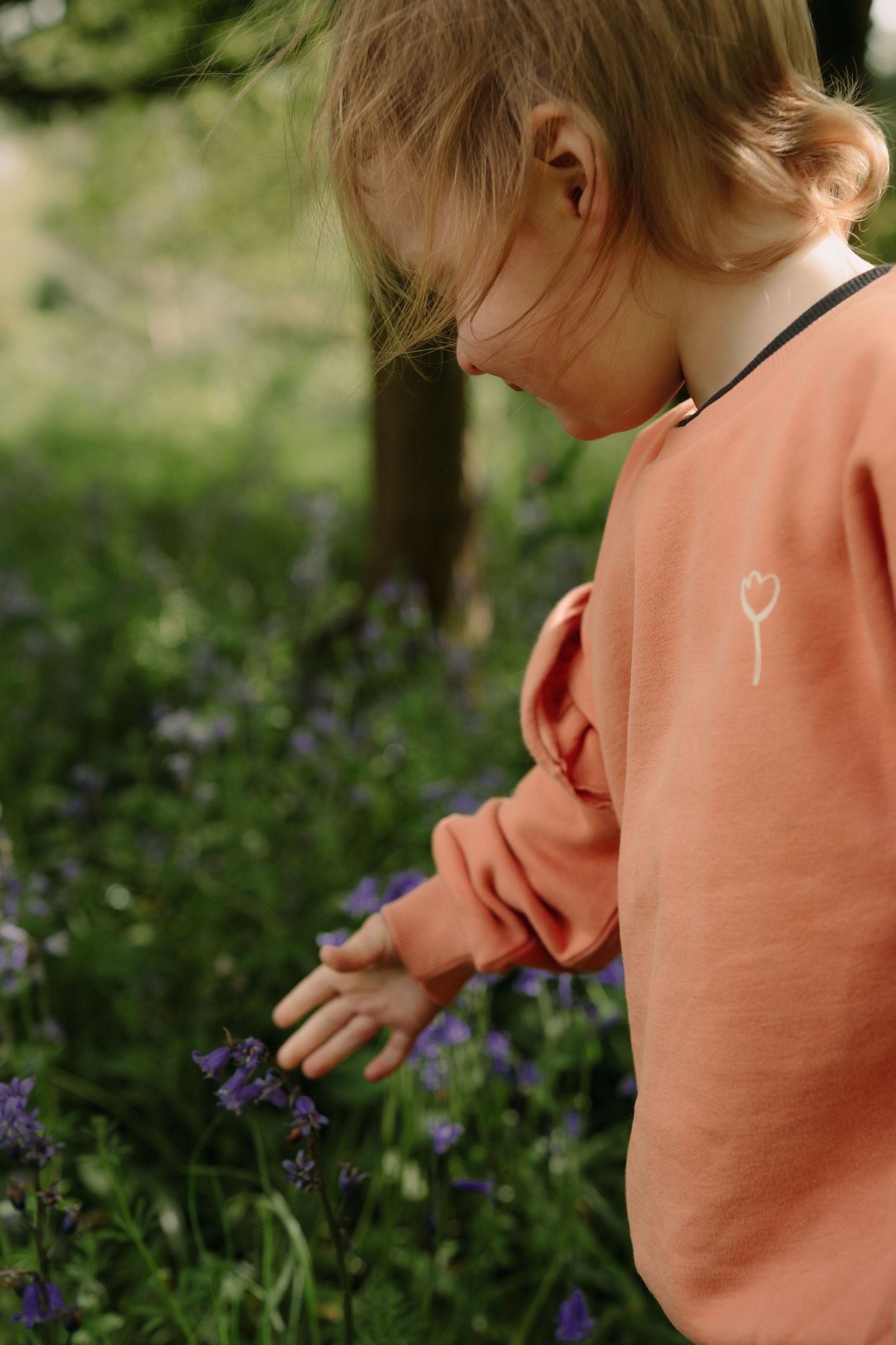 A child in a peach-colored sweater bends down to examine purple wildflowers growing in a garden.