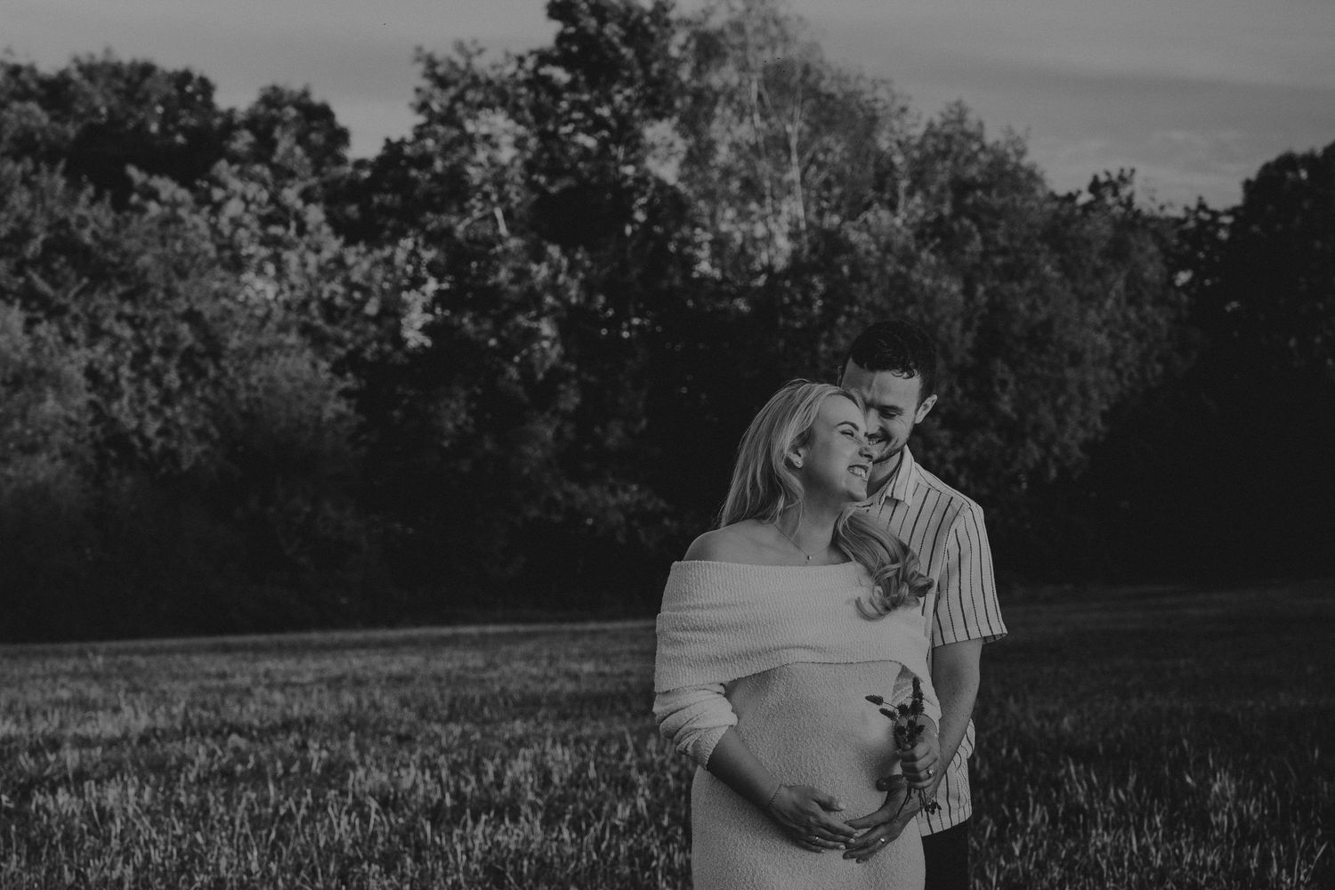 Black and white maternity photo of a couple embracing in a field surrounded by lush trees at dusk.