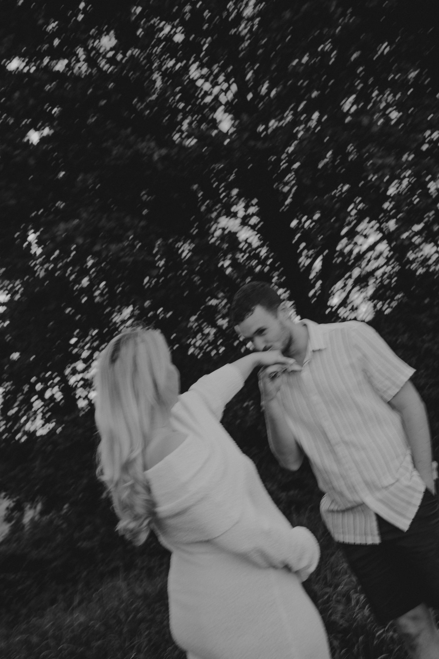 Black and white photo of a couple dancing outdoors, man kissing woman's hand near trees.