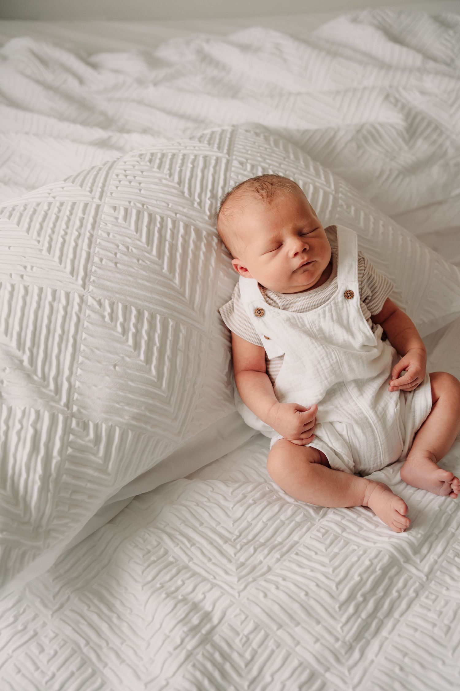 Newborn baby wearing white outfit lying on textured white bedding.