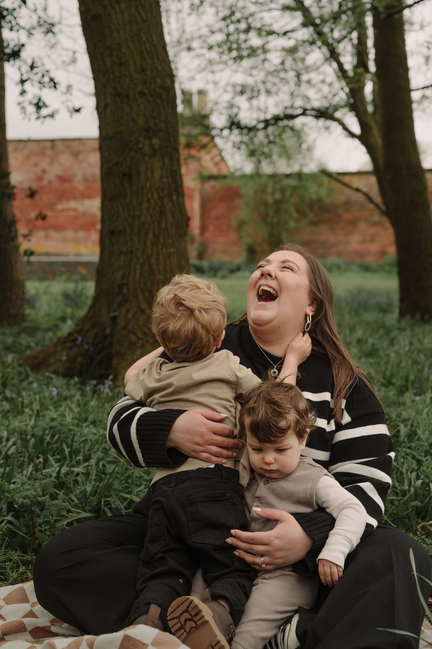 A joyful outdoor family moment captured on a blanket under tall trees with a brick wall in the background.