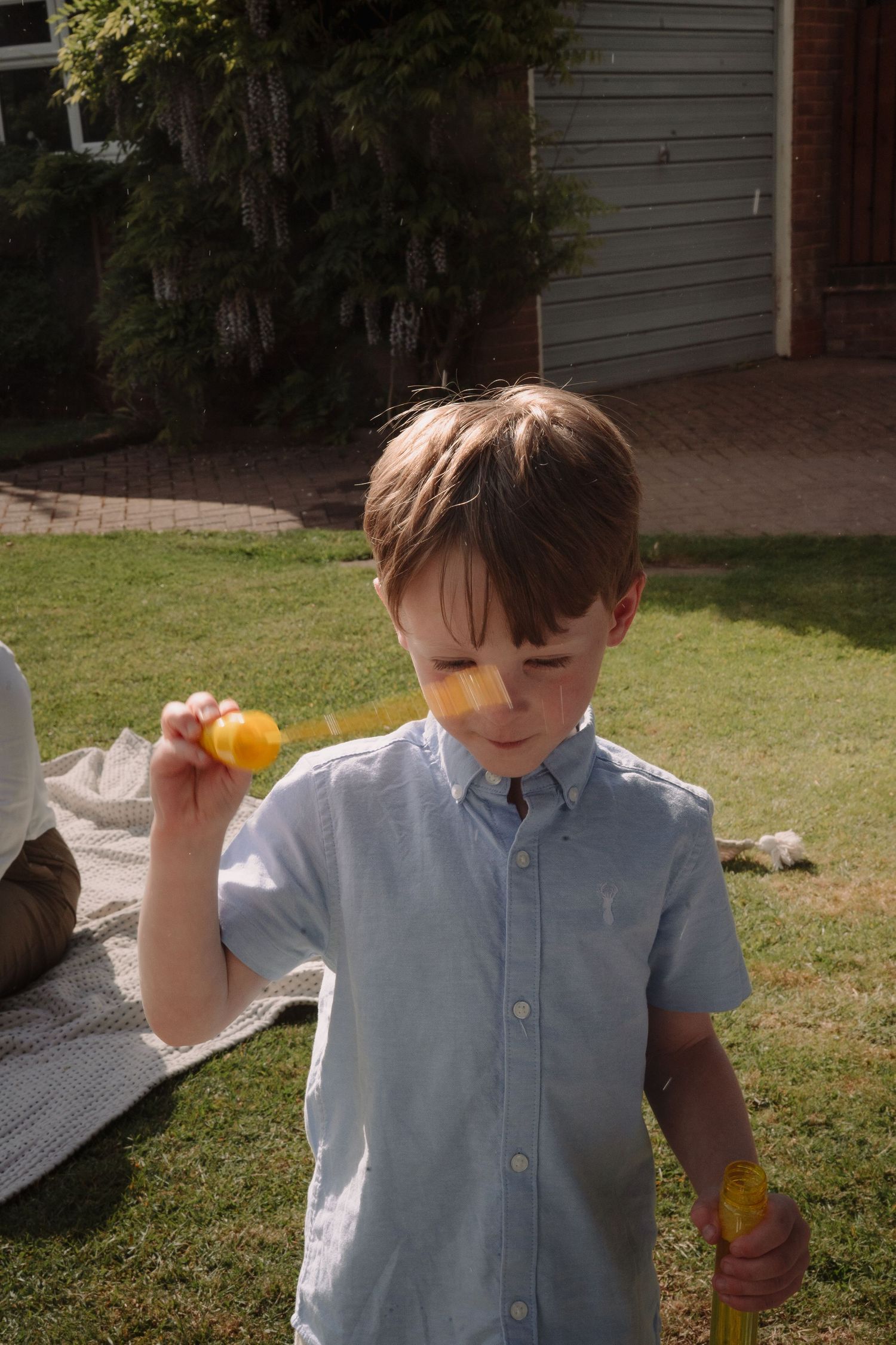 Young boy in light blue shirt holding a yellow Easter egg in a sunny backyard garden.