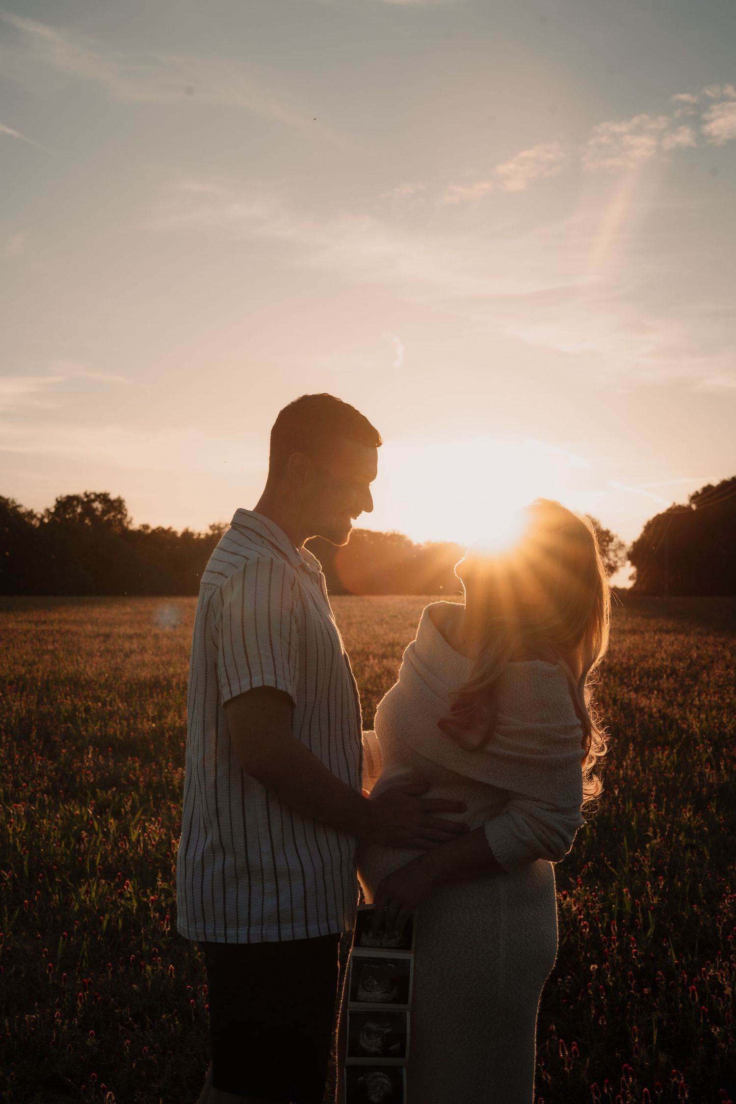 Couple sharing a tender moment in a golden sunset field, woman holding ultrasound photos during a maternity announcement shoot.