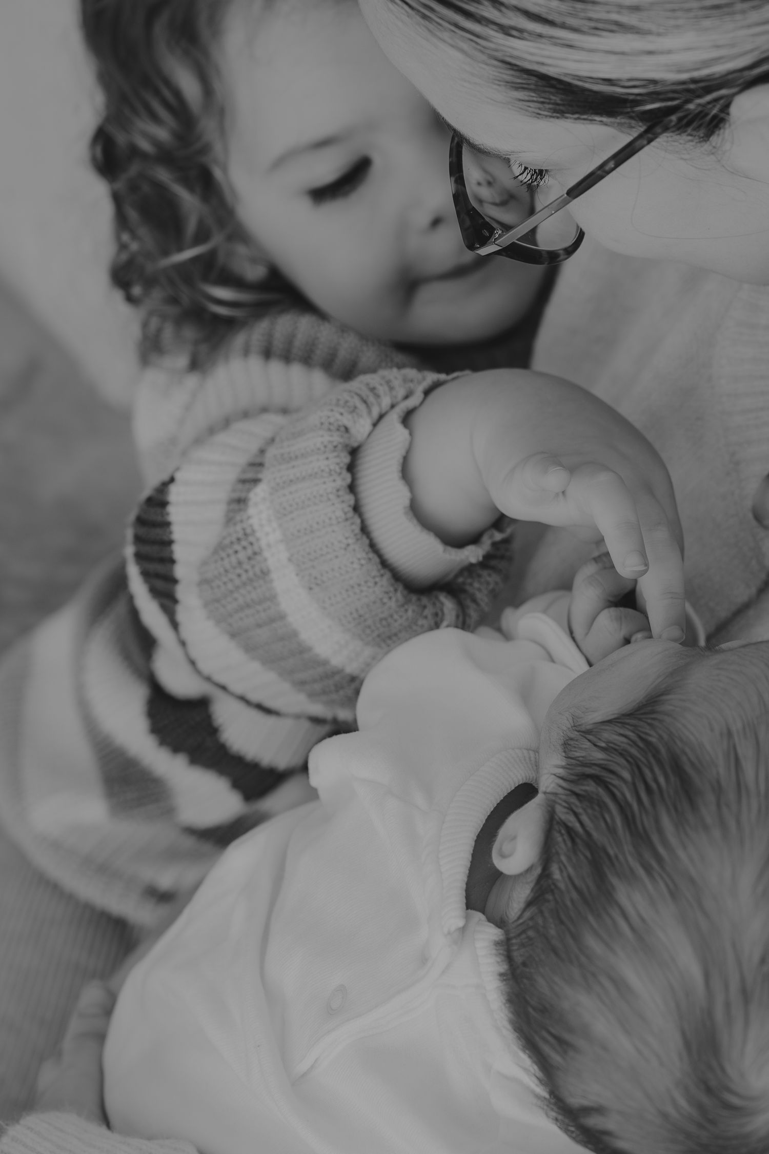 A tender black and white photograph showing a close moment between siblings with a baby in a striped sweater.