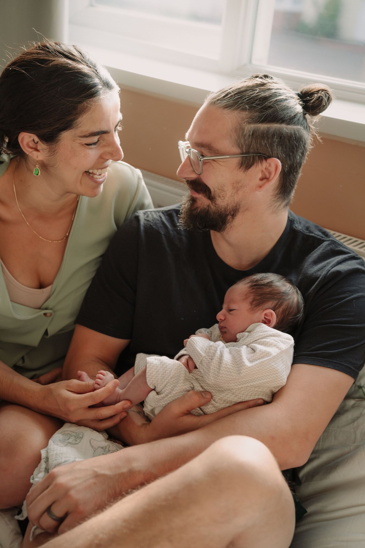 Parents snuggle with newborn baby on couch in series of tender moments captured in natural light.