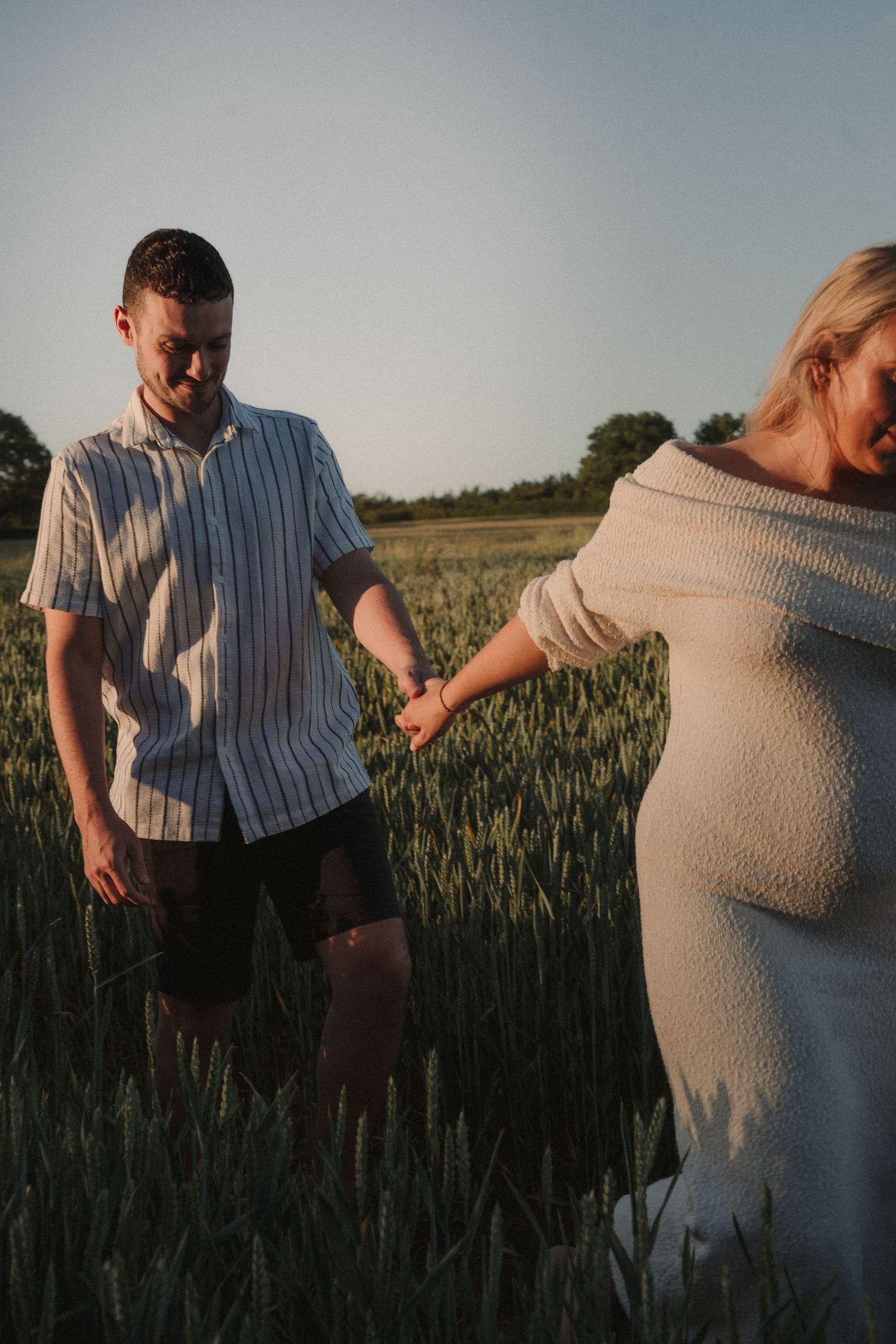 Couple holding hands in a golden wheat field at sunset, woman in white off-shoulder dress showing baby bump.