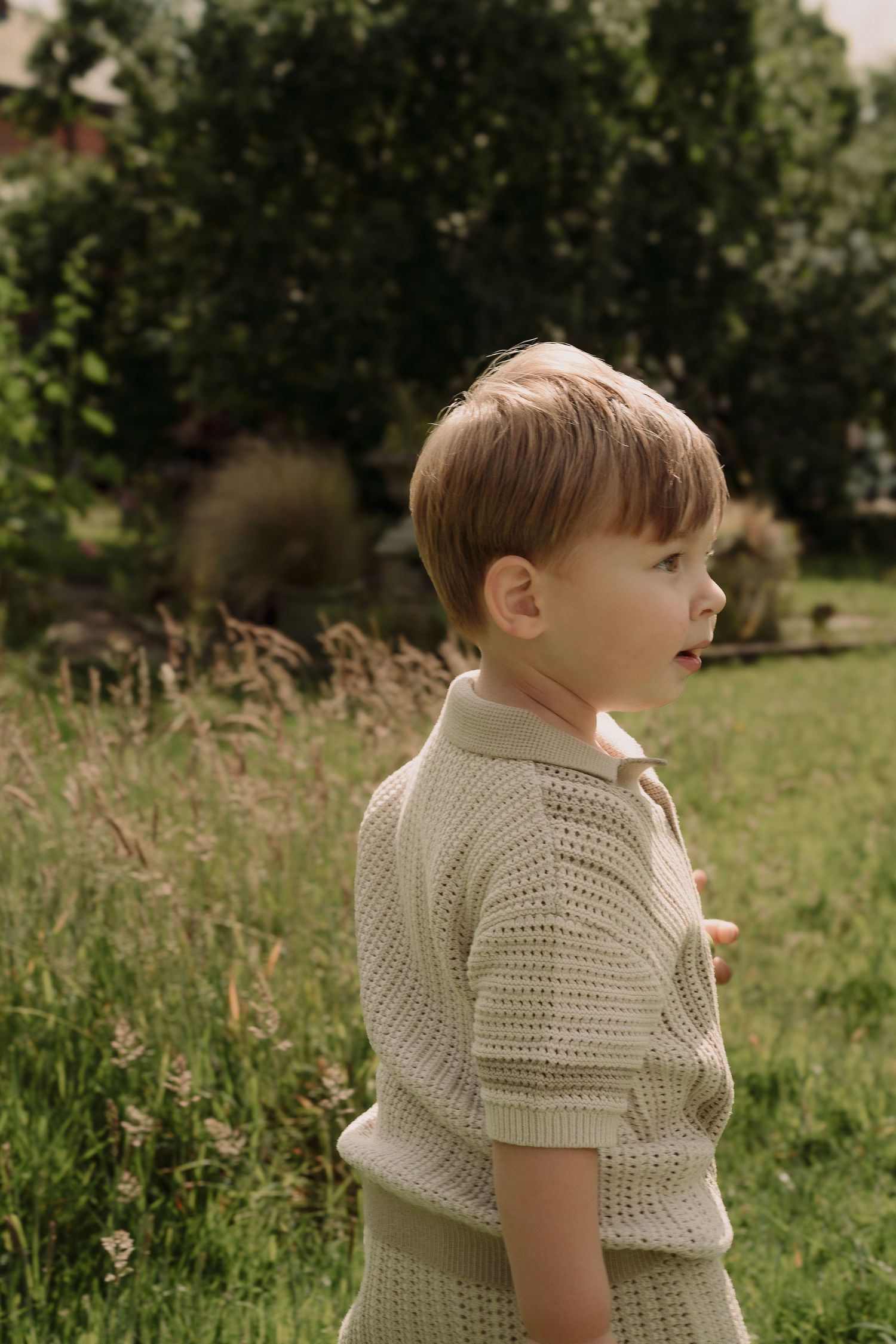 A child wearing a white knitted sweater stands in a grassy meadow on a sunny day by Family photographer in Tamworth.