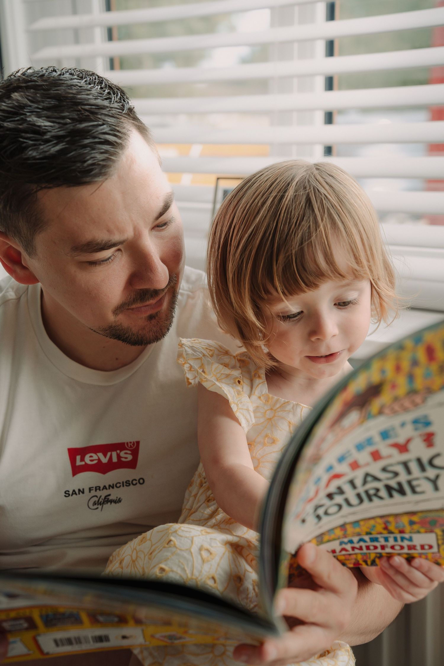 A parent and young child read a colorful storybook together while sitting by a window with venetian blinds.
