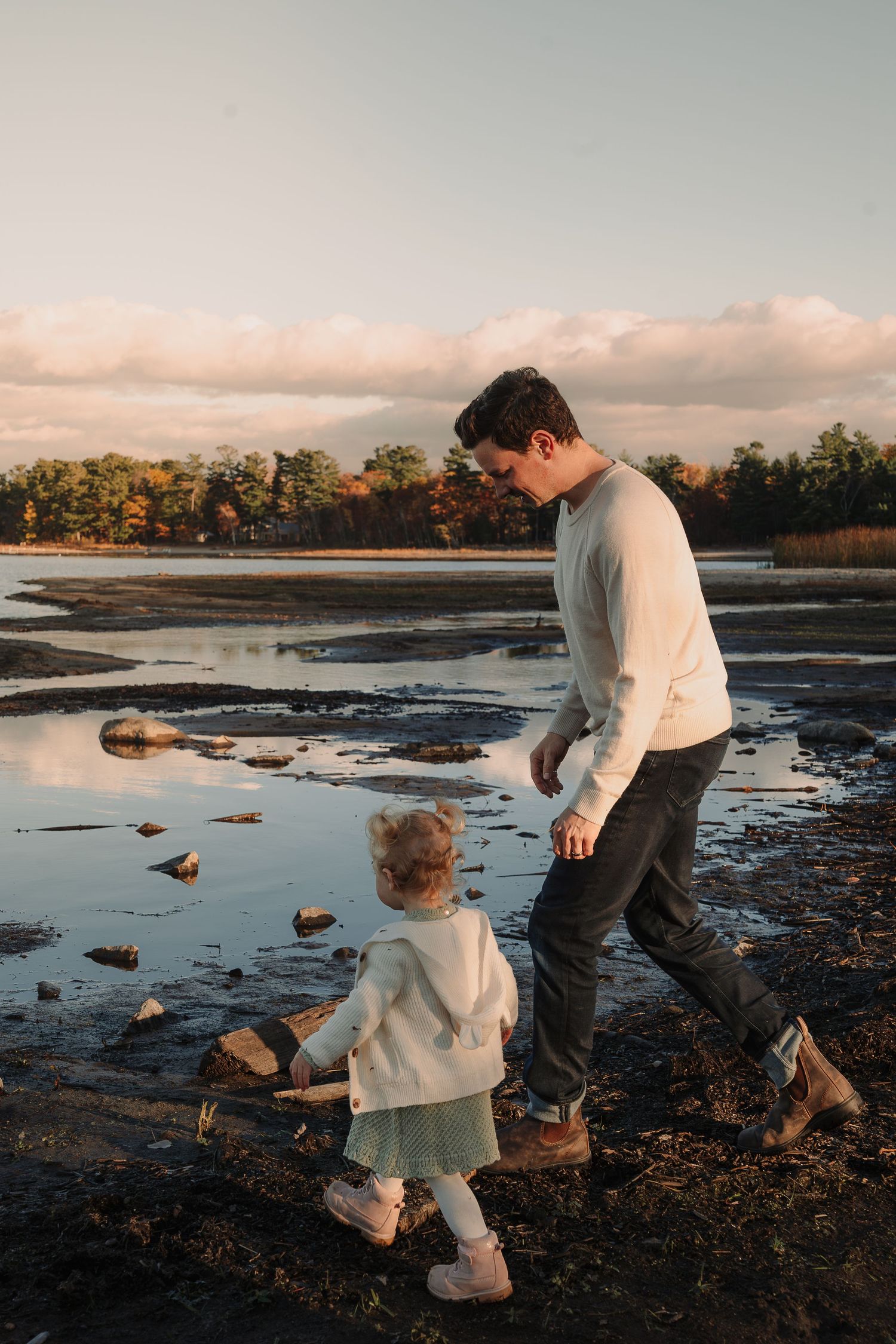 Parent and child exploring a muddy shoreline at sunset.