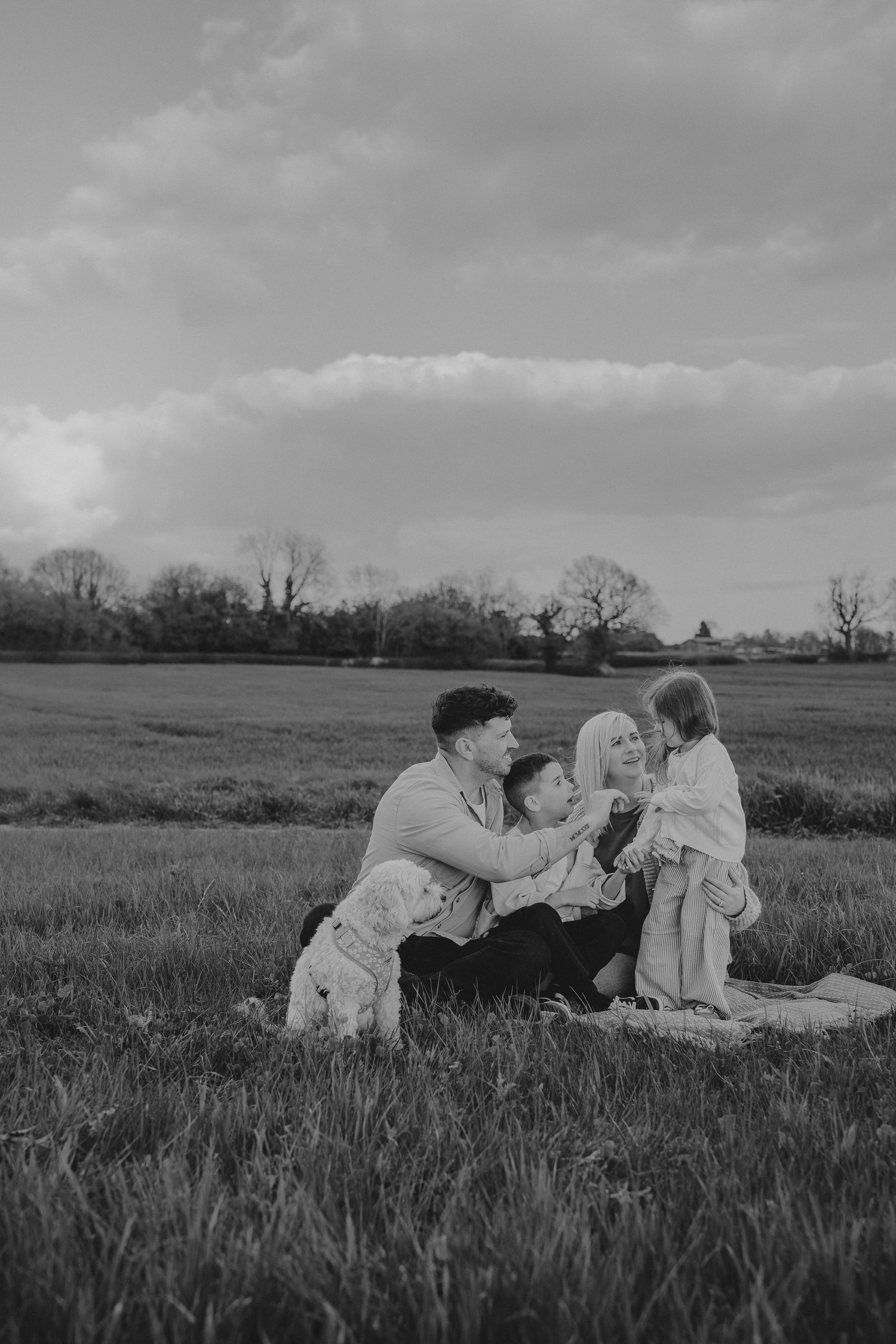 Black and white family photo of parents and two children playing together on a blanket in a grassy field.