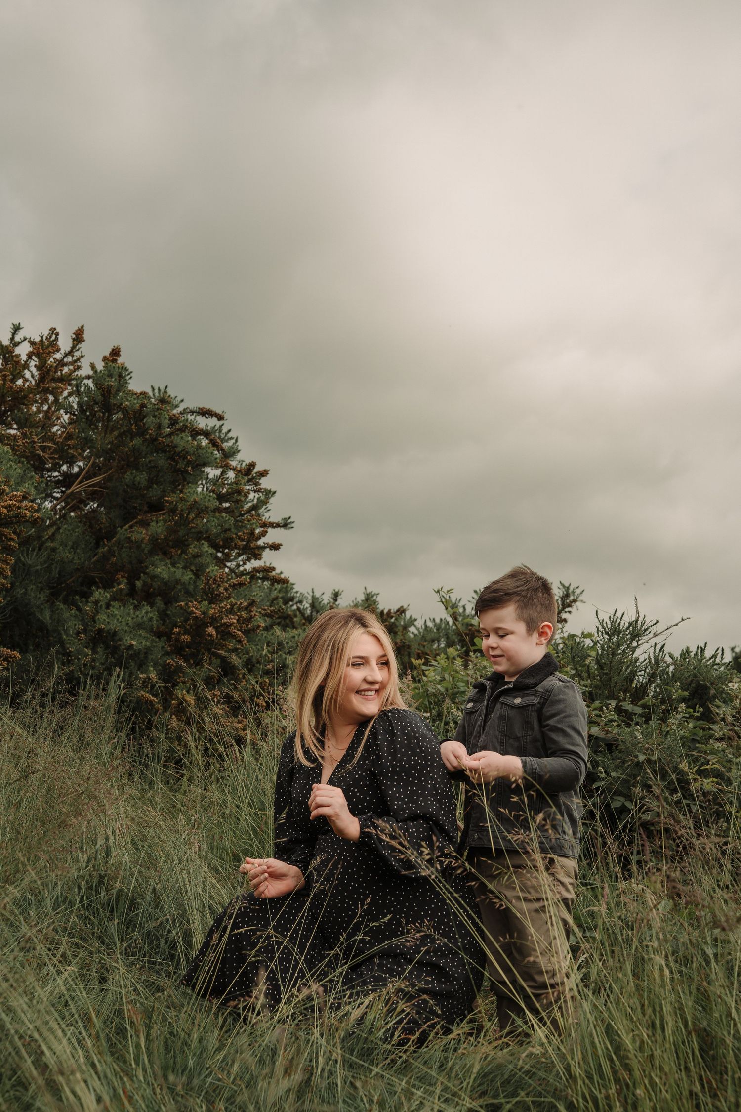 Two people sit together in tall grass on an overcast day, sharing a playful moment in nature.