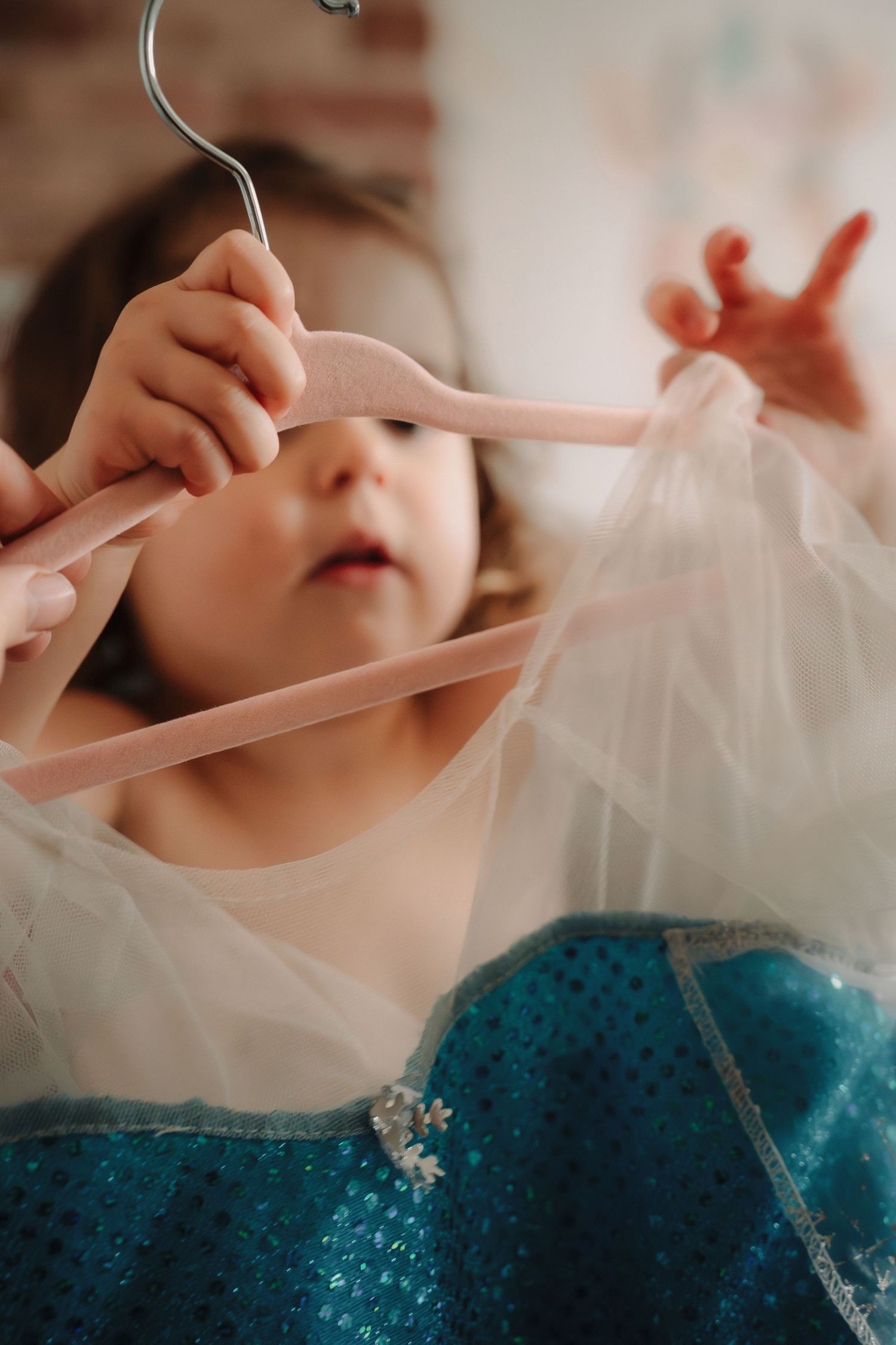 Small child playing with delicate pink hangers and sheer fabric near a sparkly blue dress.