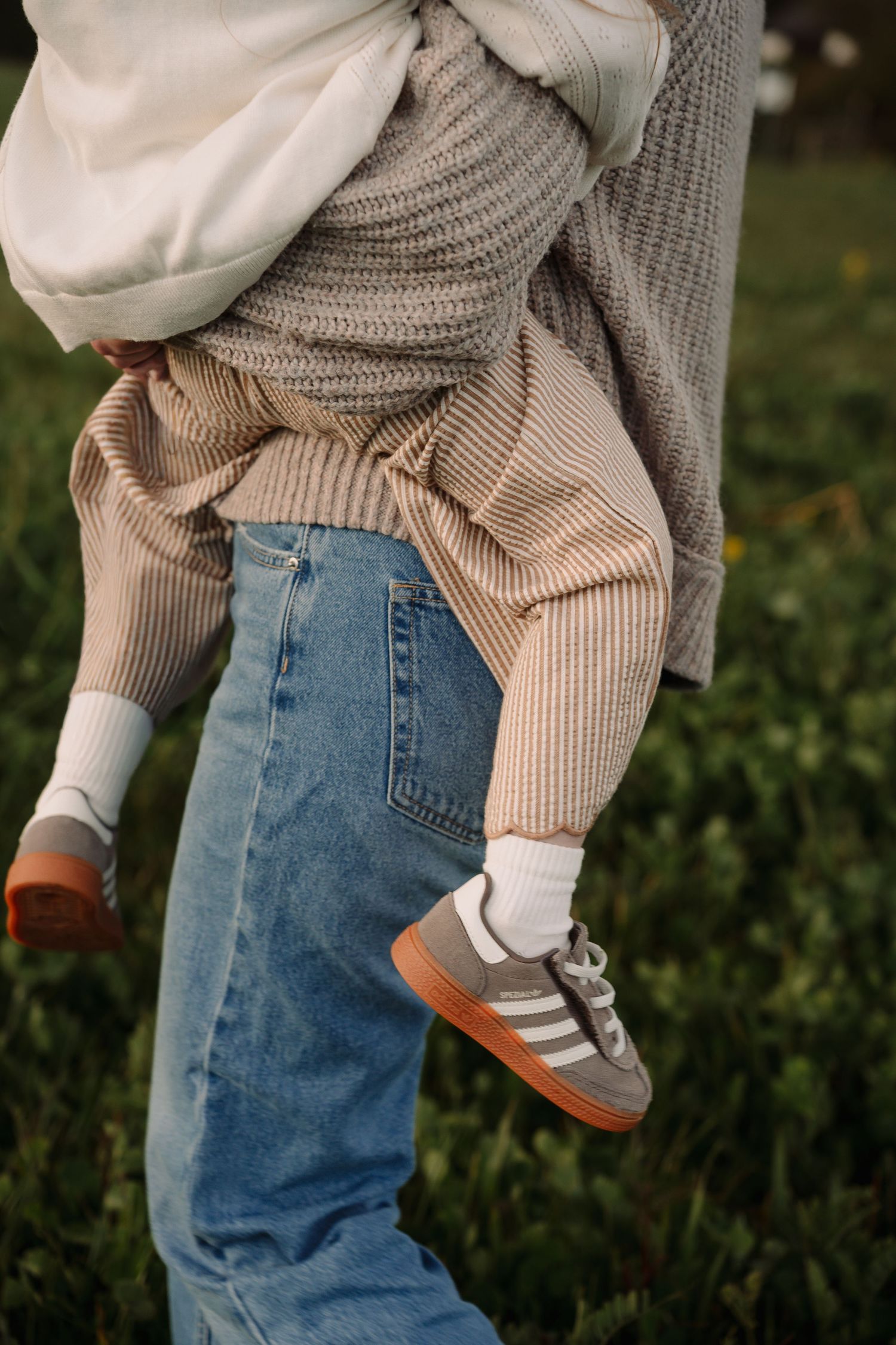 Parent in blue jeans and Adidas Samba sneakers carrying a small child over their shoulder outdoors.