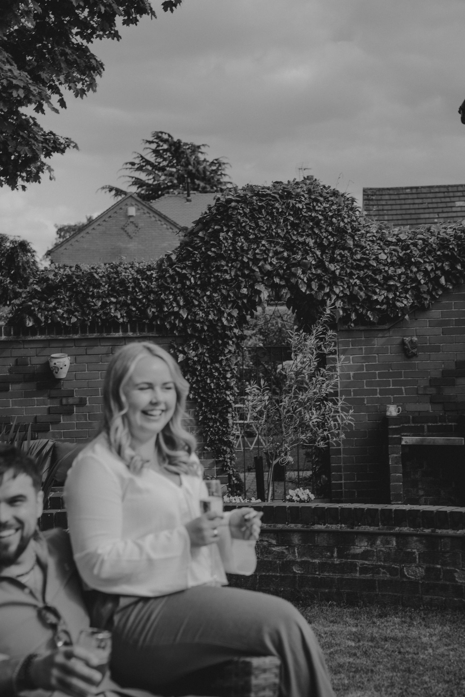 Black and white photo of a laughing blonde woman holding a drink outdoors, sitting by a brick wall covered in ivy.