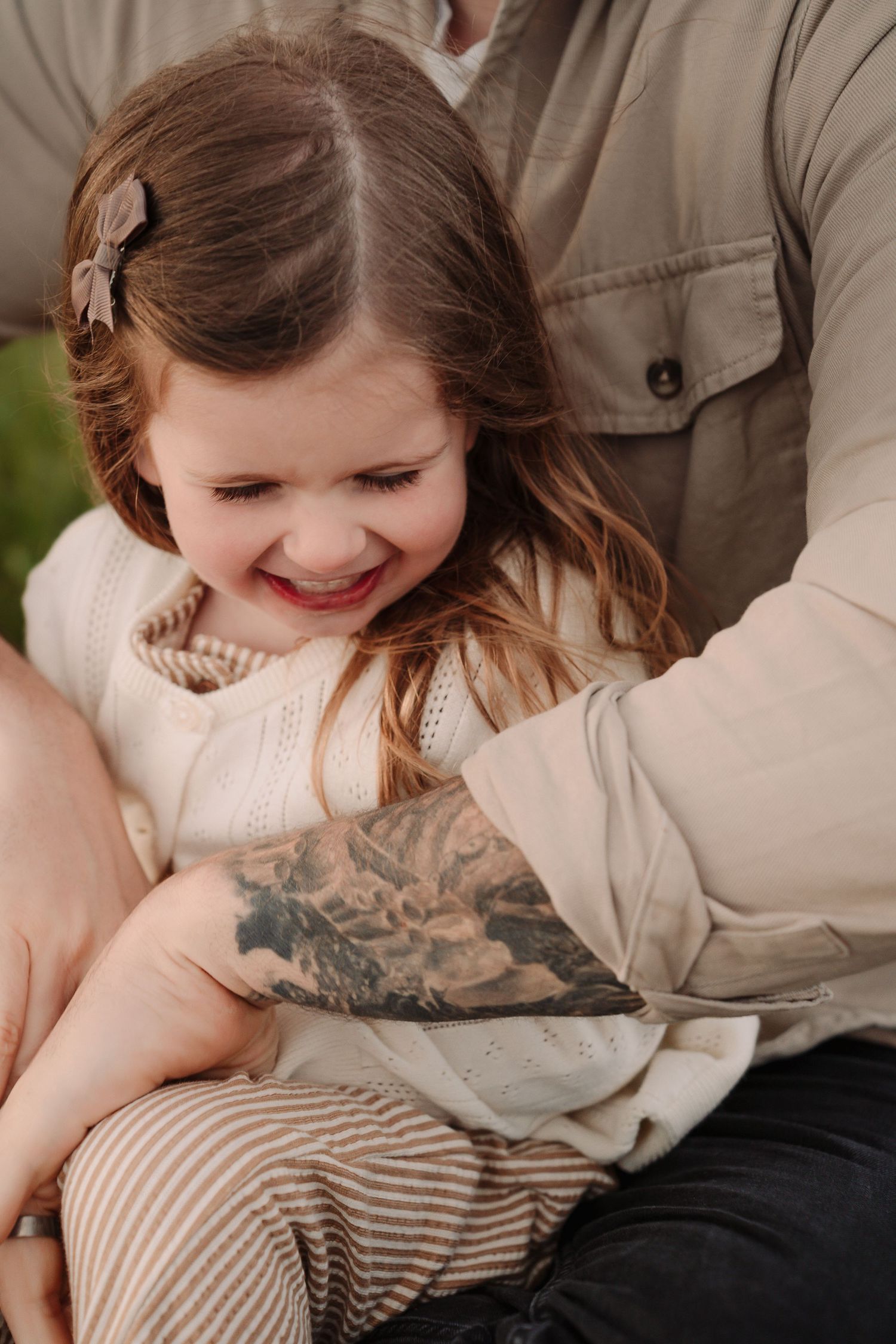 A smiling little girl in a cream knit dress sits on her tattooed fathers lap outdoors.