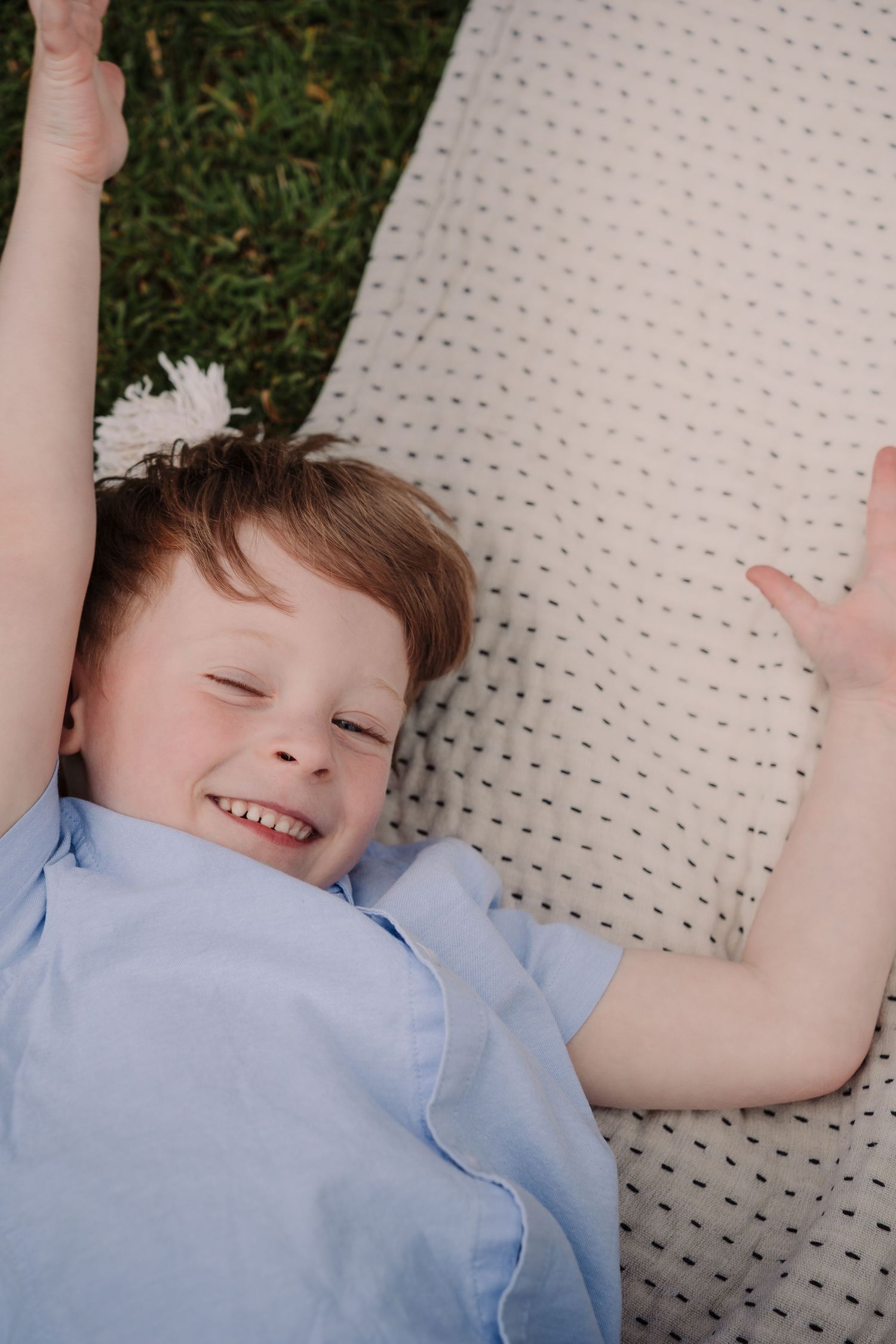 A happy young boy lying on a blanket outdoors, smiling with arms raised, wearing a light blue shirt.