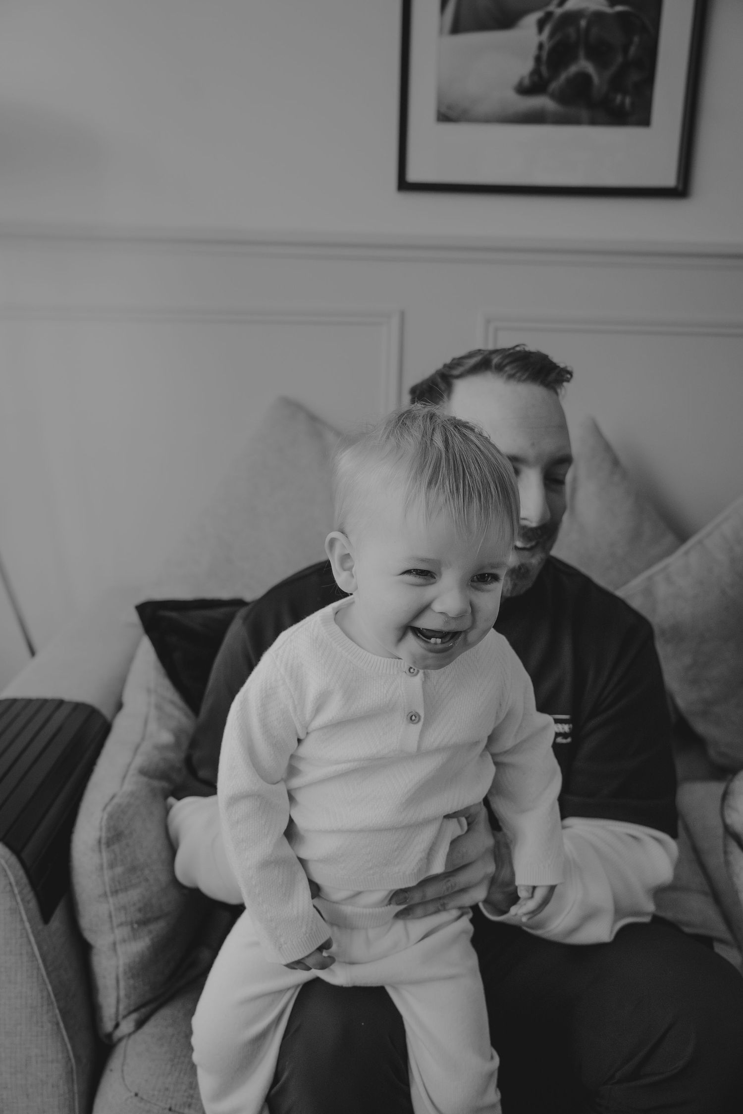 Black and white photo of a laughing baby in white pajamas sitting on a man's lap on a couch.