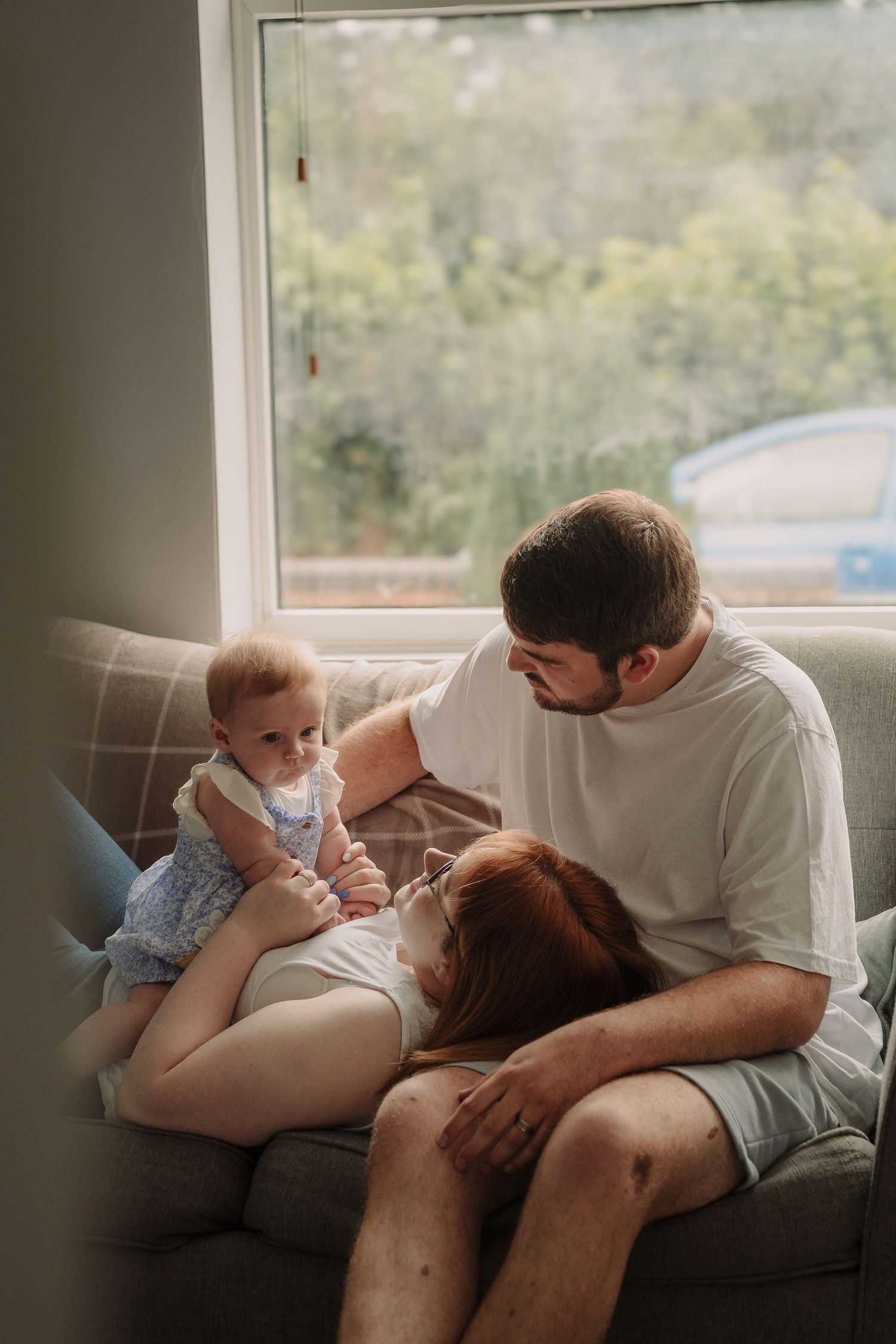 A family relaxing together on a gray couch by a large window with natural lighting.