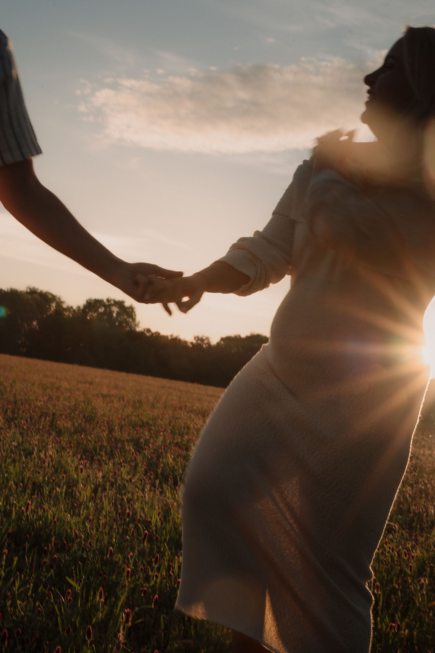 Couple holding hands in a golden sunset field, woman in white dress with sun flare, romantic outdoor maternity photo.