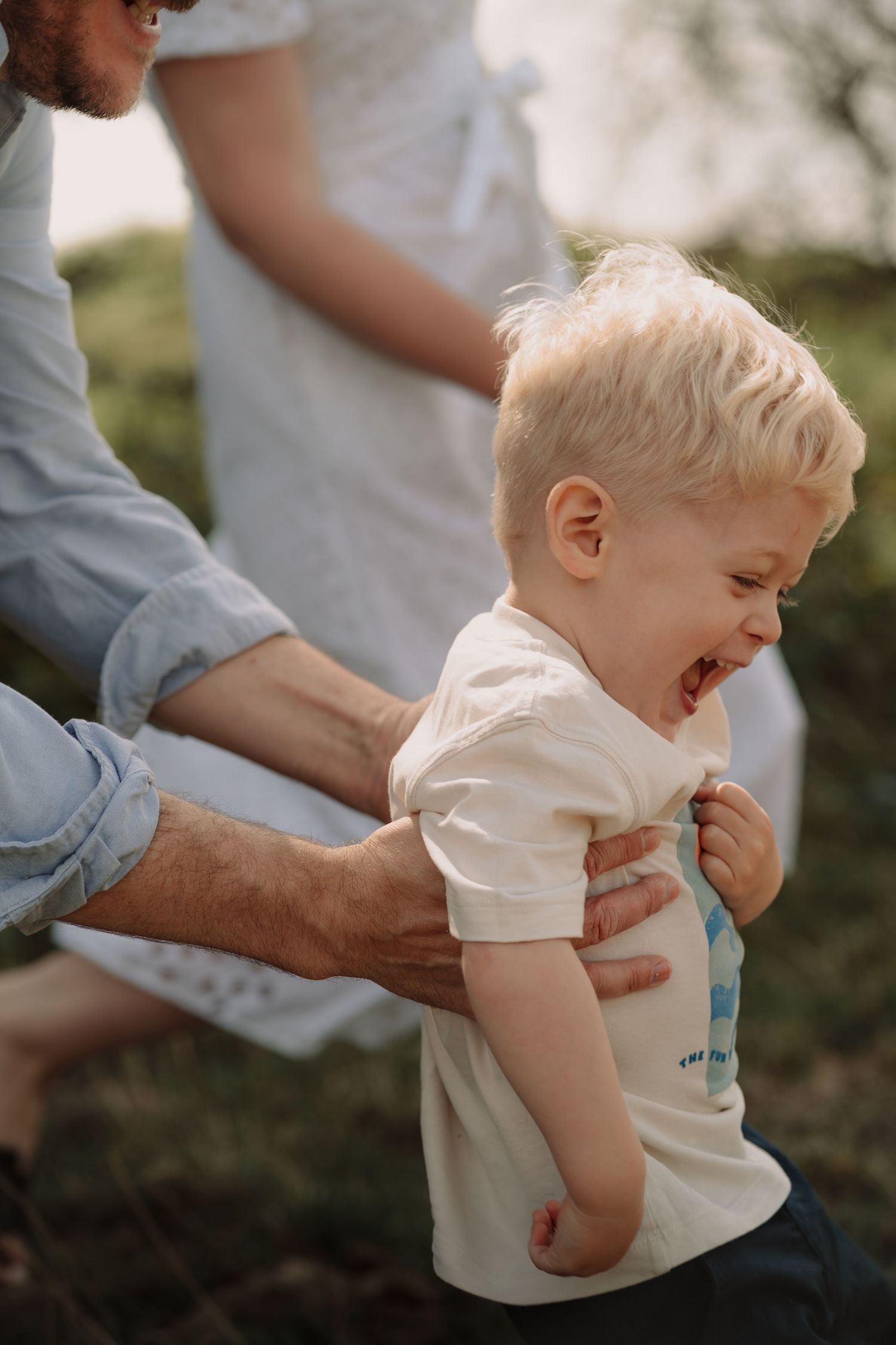 Playful sequence of a giggling toddler in a white shirt being tickled outdoors.