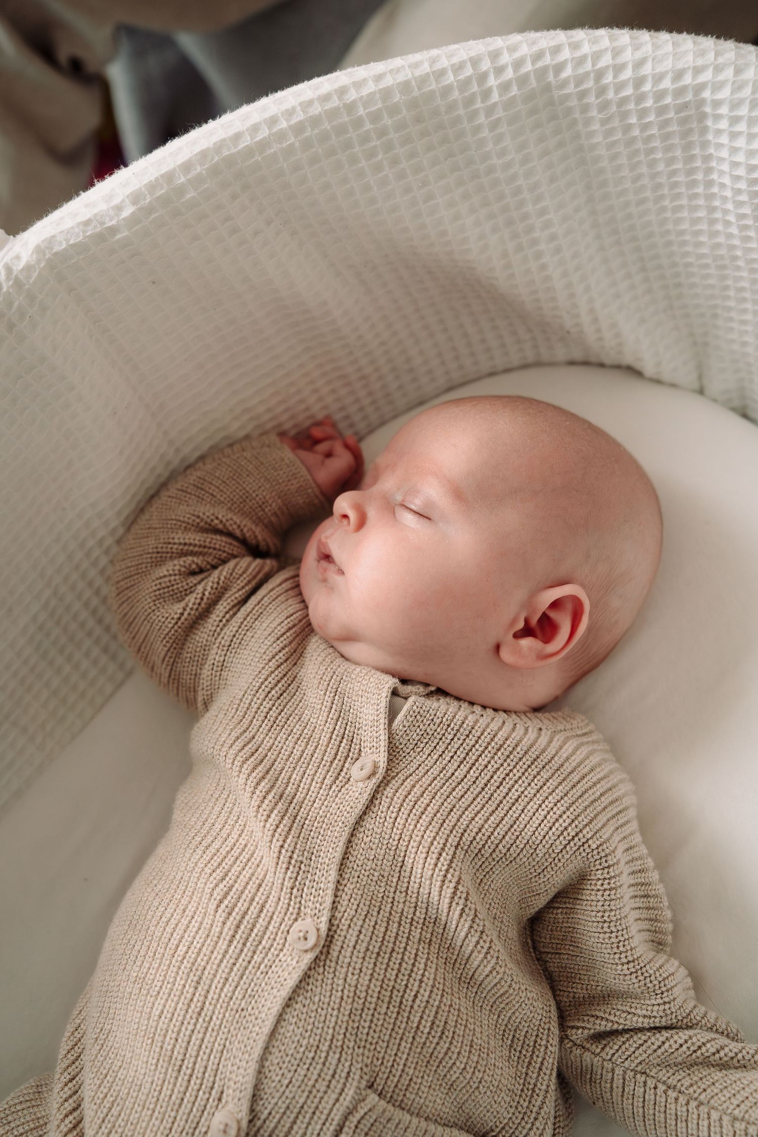 Sleeping infant wearing beige knit cardigan rests on white textured surface.