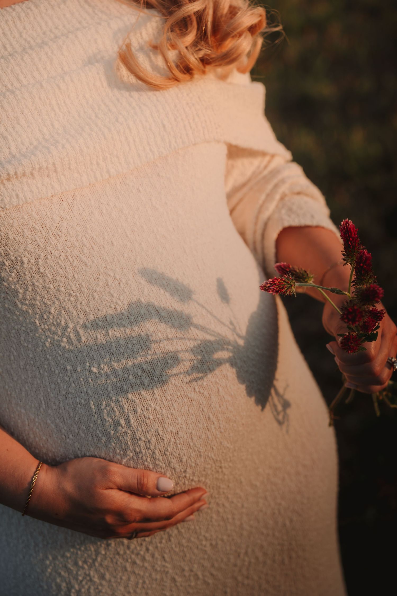 Pregnant woman in white knit dress holding red wildflowers, with shadow cast on belly in warm golden light.