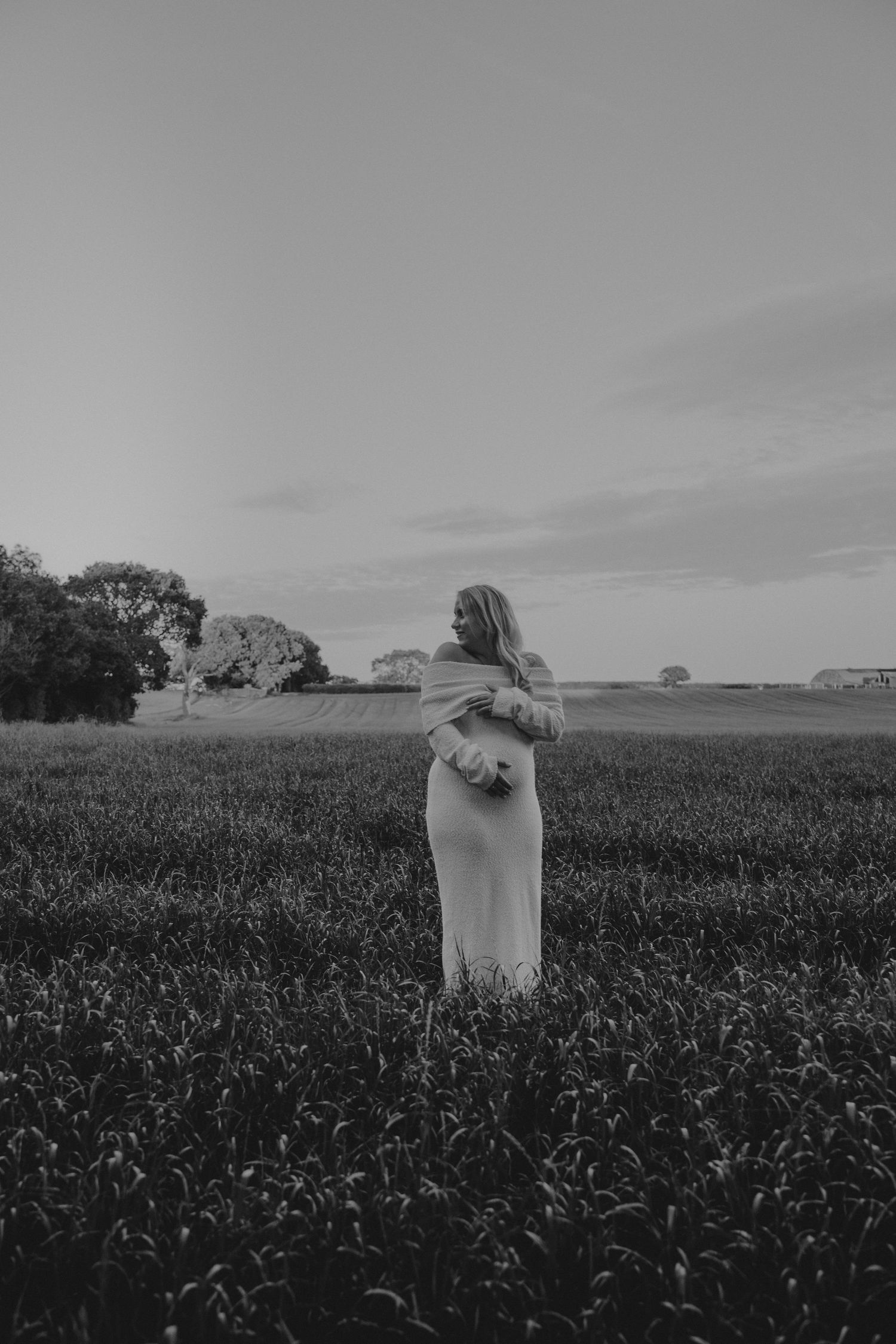 Black and white photo of a woman in a field at dusk, wearing a long dress, surrounded by lush grass and trees.