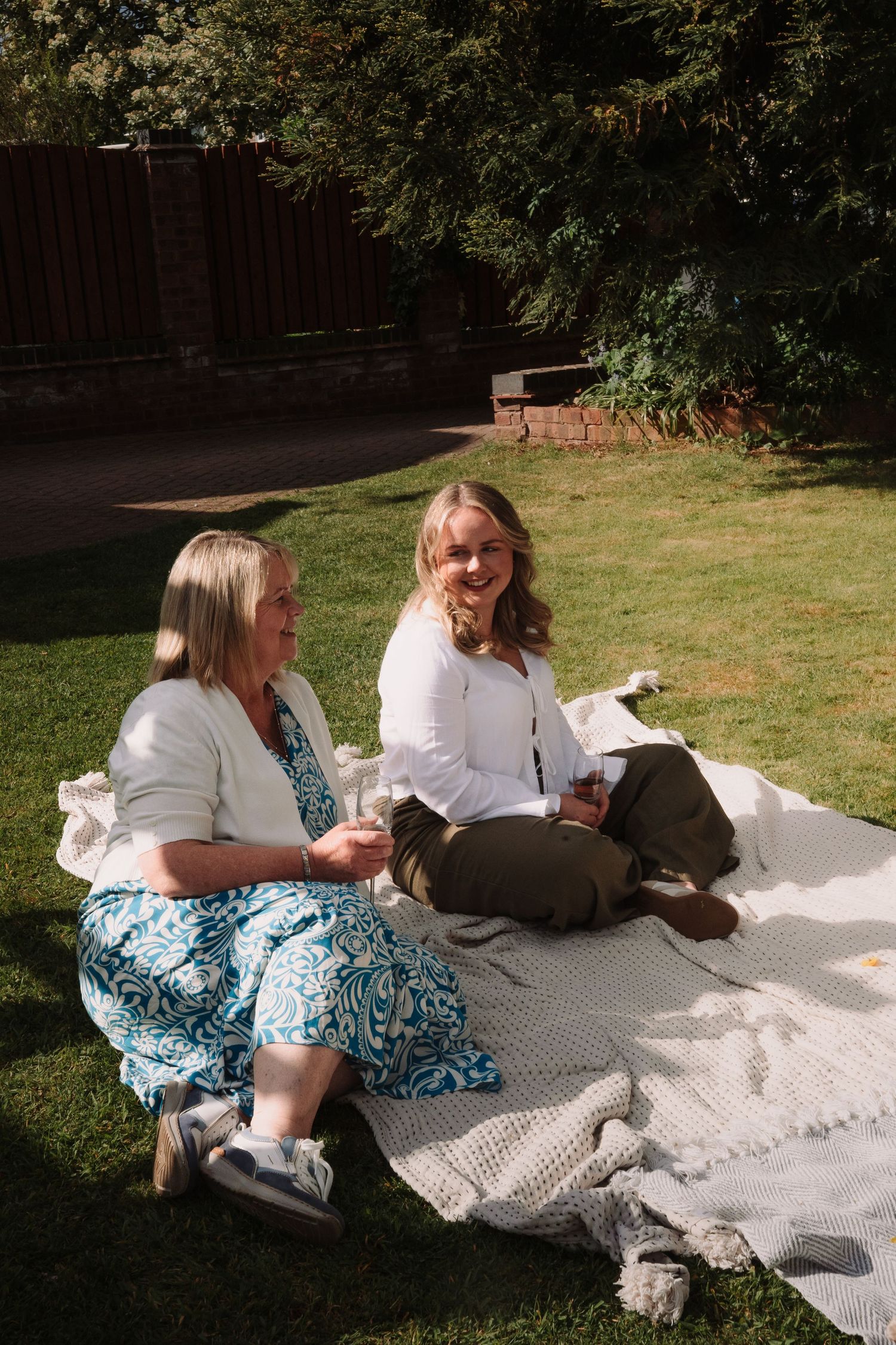 Two women sitting on a blanket in a sunny garden, smiling and relaxing outdoors with a small dog.