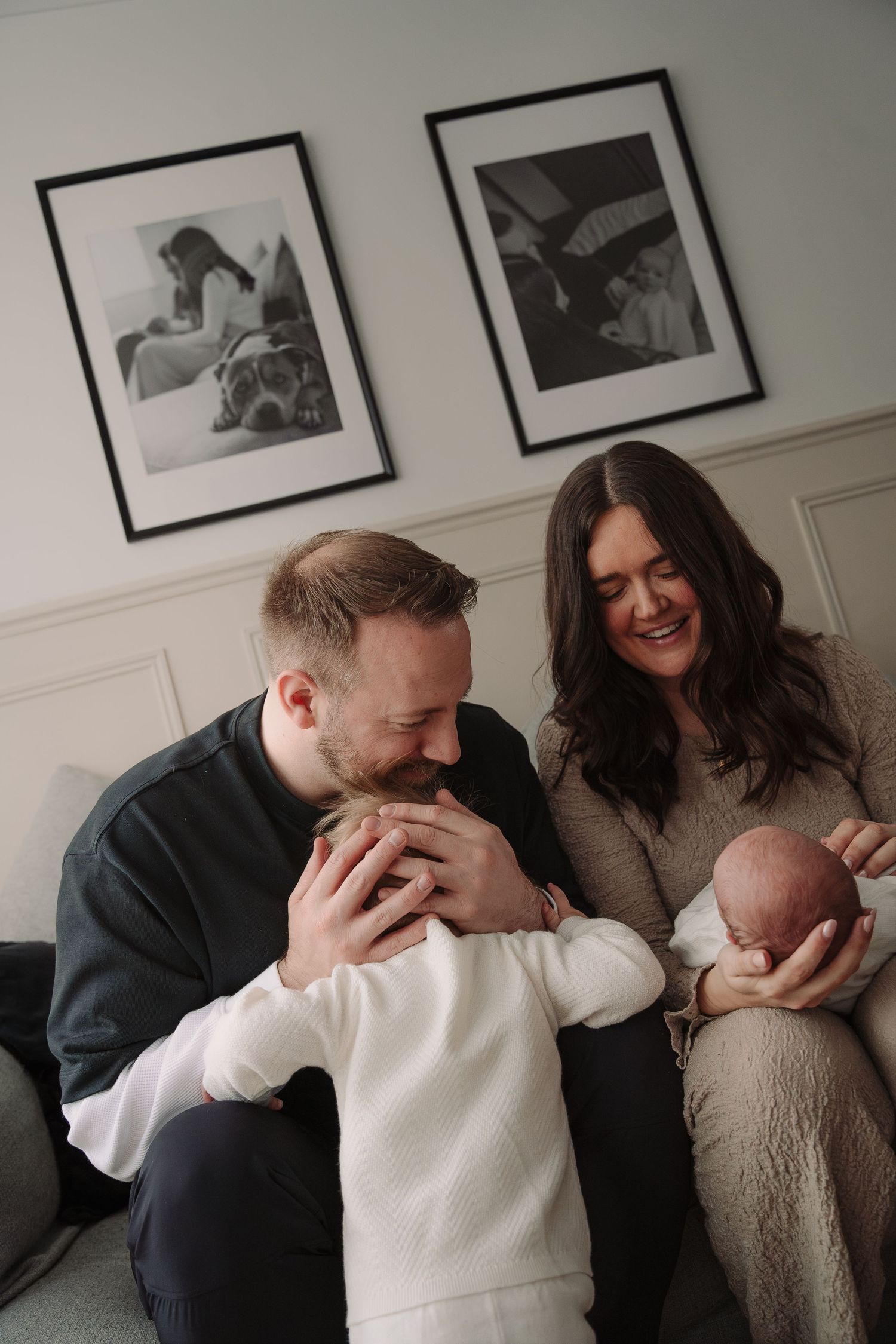 Happy family with newborn baby and toddler sitting on sofa, black and white framed photos on wall behind them.