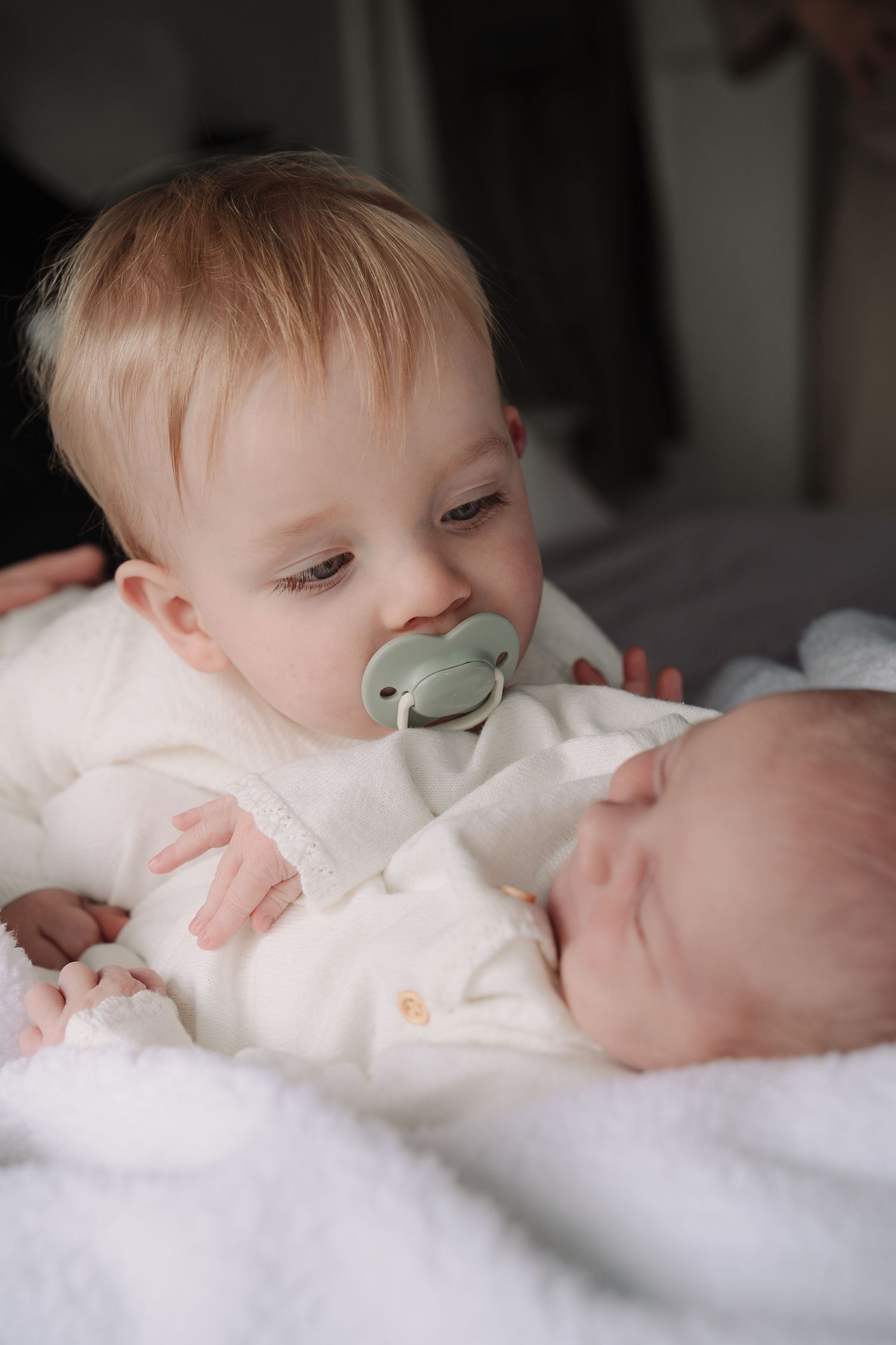 Toddler with pacifier in white outfit gently holds sleeping newborn baby sibling on soft white blanket.