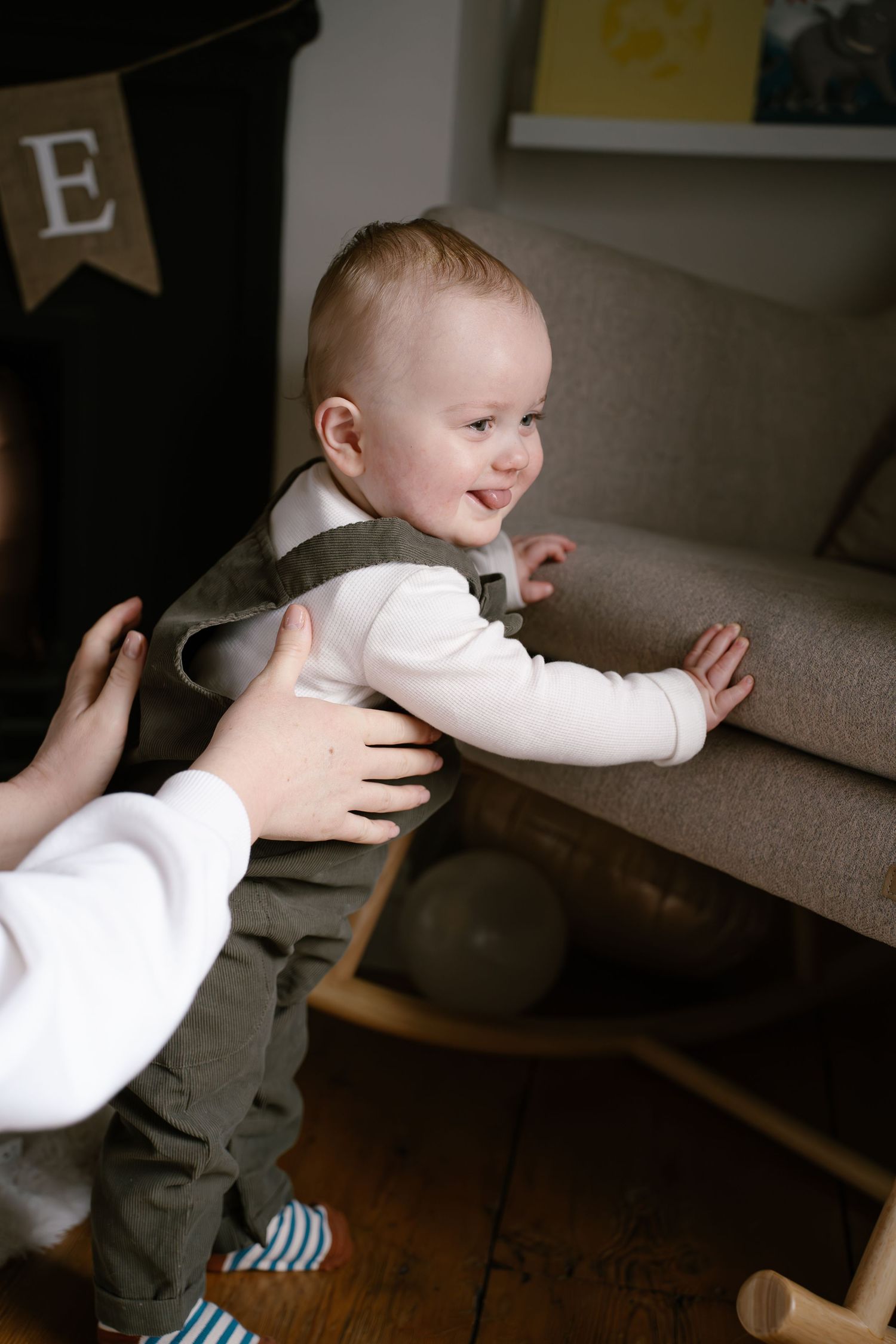 Baby wearing white shirt and overalls smiles while being supported near a gray couch in a living room.