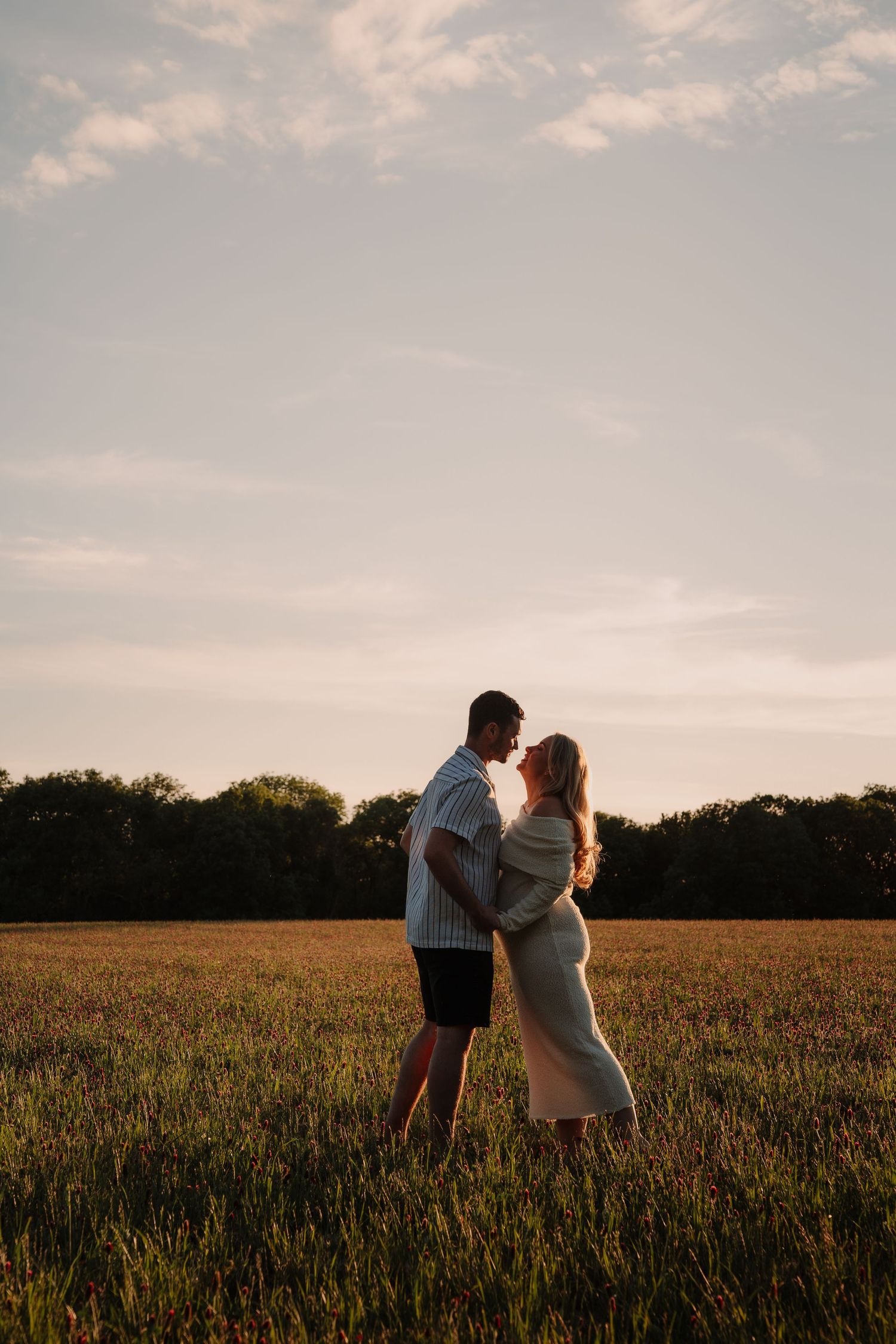 Couple embracing in golden field at sunset, pregnant woman in dress with partner, romantic maternity photo outdoors.