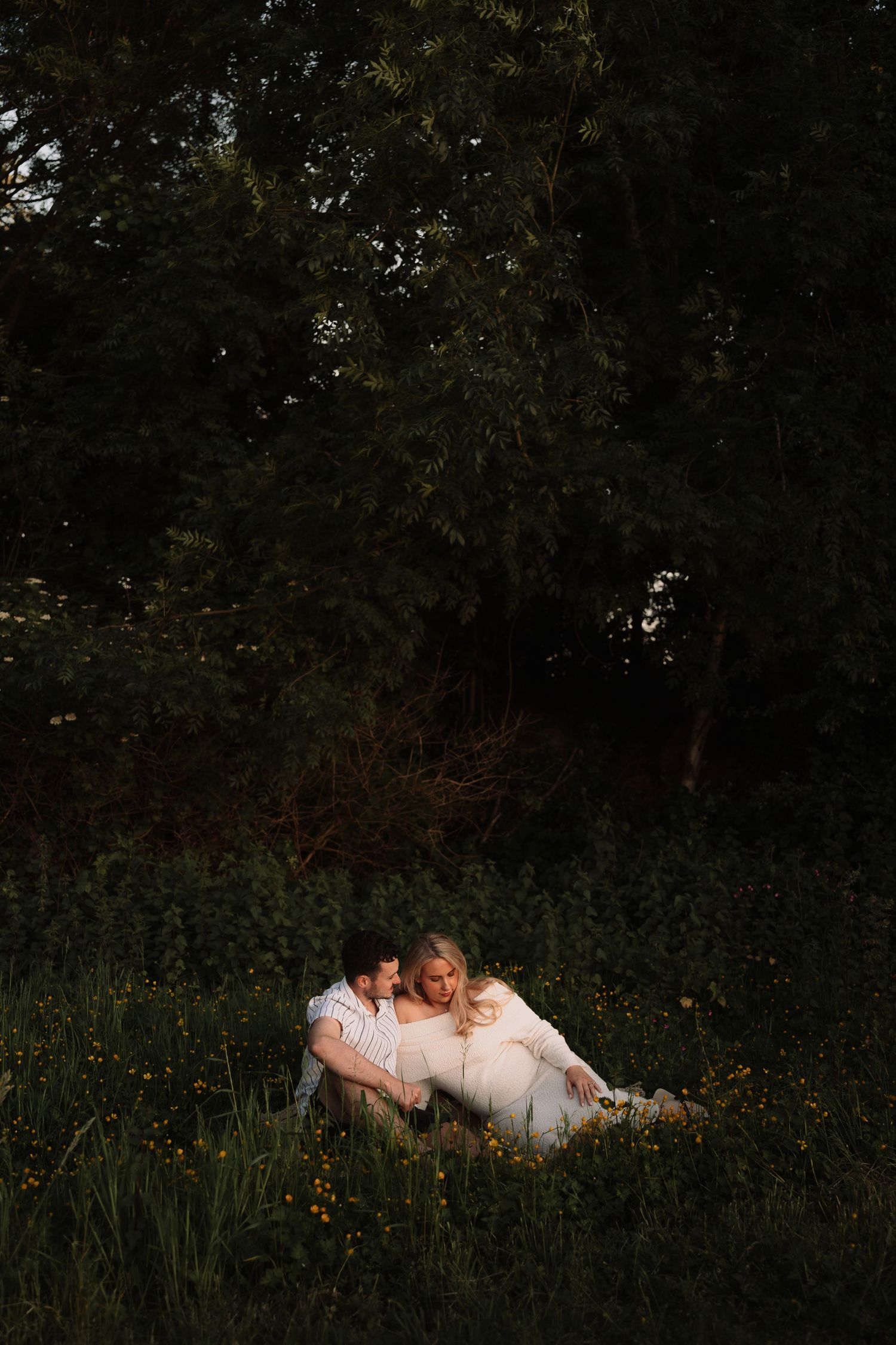 Couple sitting together in a lush green meadow at golden hour, surrounded by wildflowers and trees.