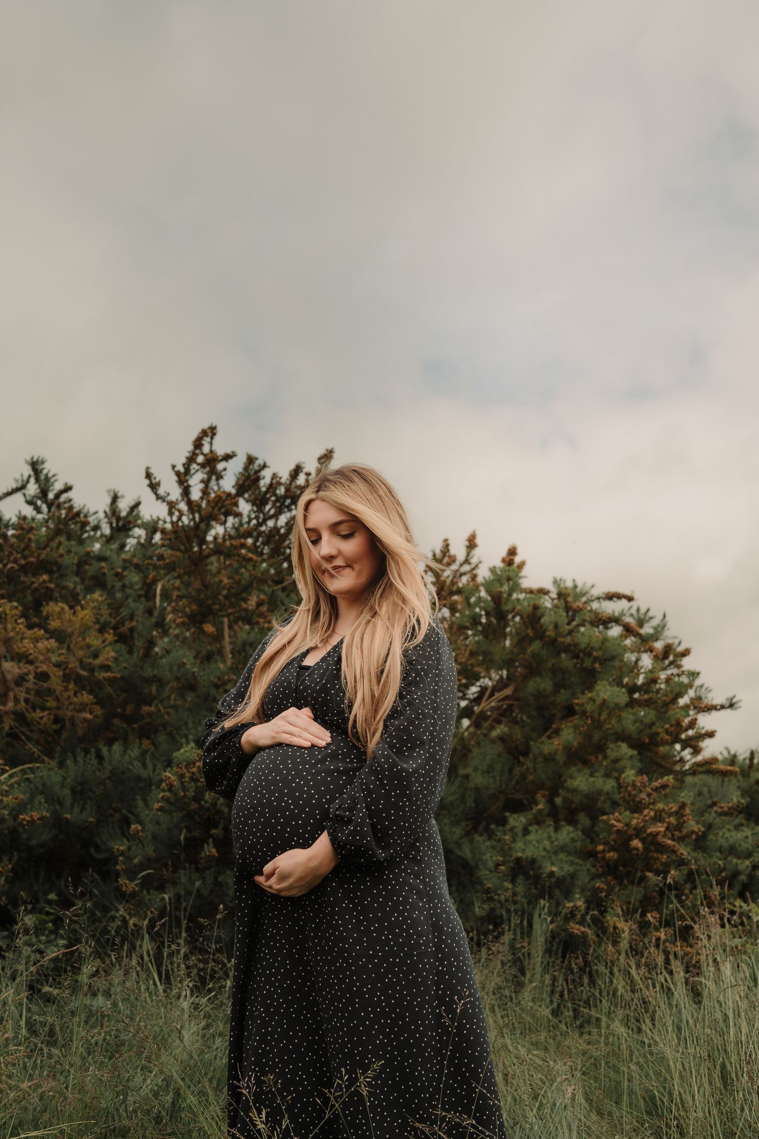 Woman in black polka dot dress poses thoughtfully in a natural outdoor setting during golden hour.