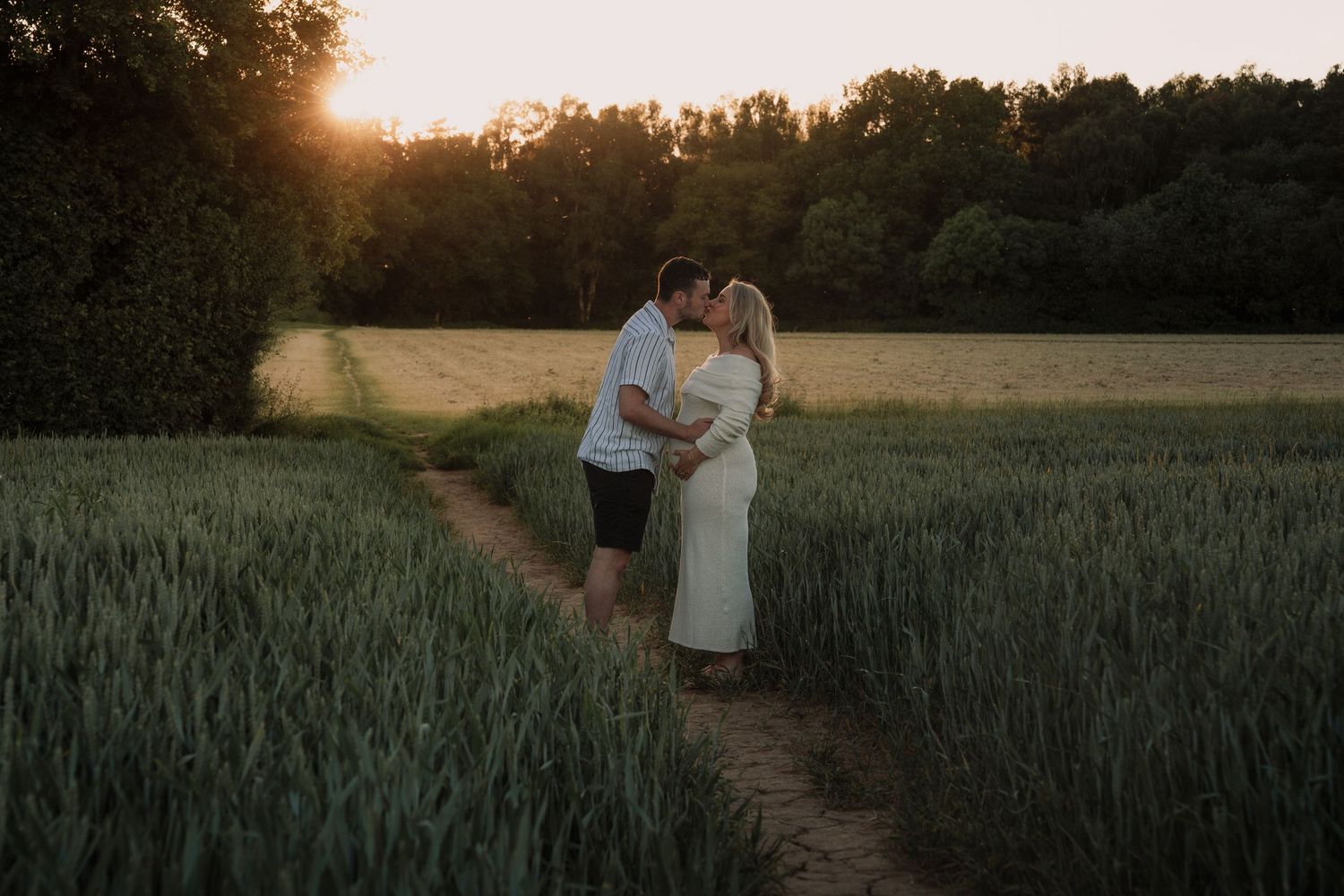 Couple kissing at sunset in a green wheat field, golden light filtering through trees in a romantic outdoor portrait.