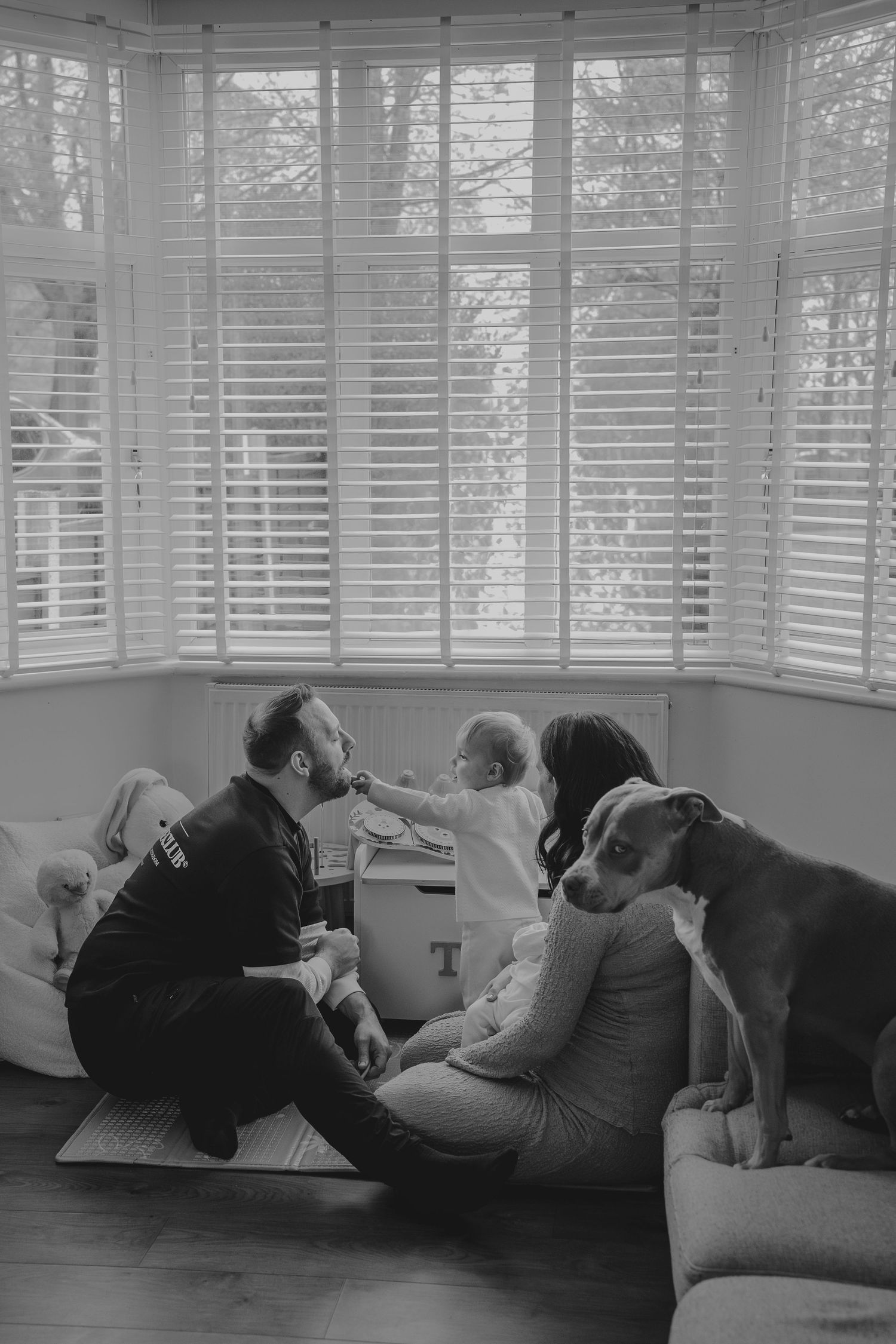 Black and white family photo with parents, baby, and dog by bay window with shutters.